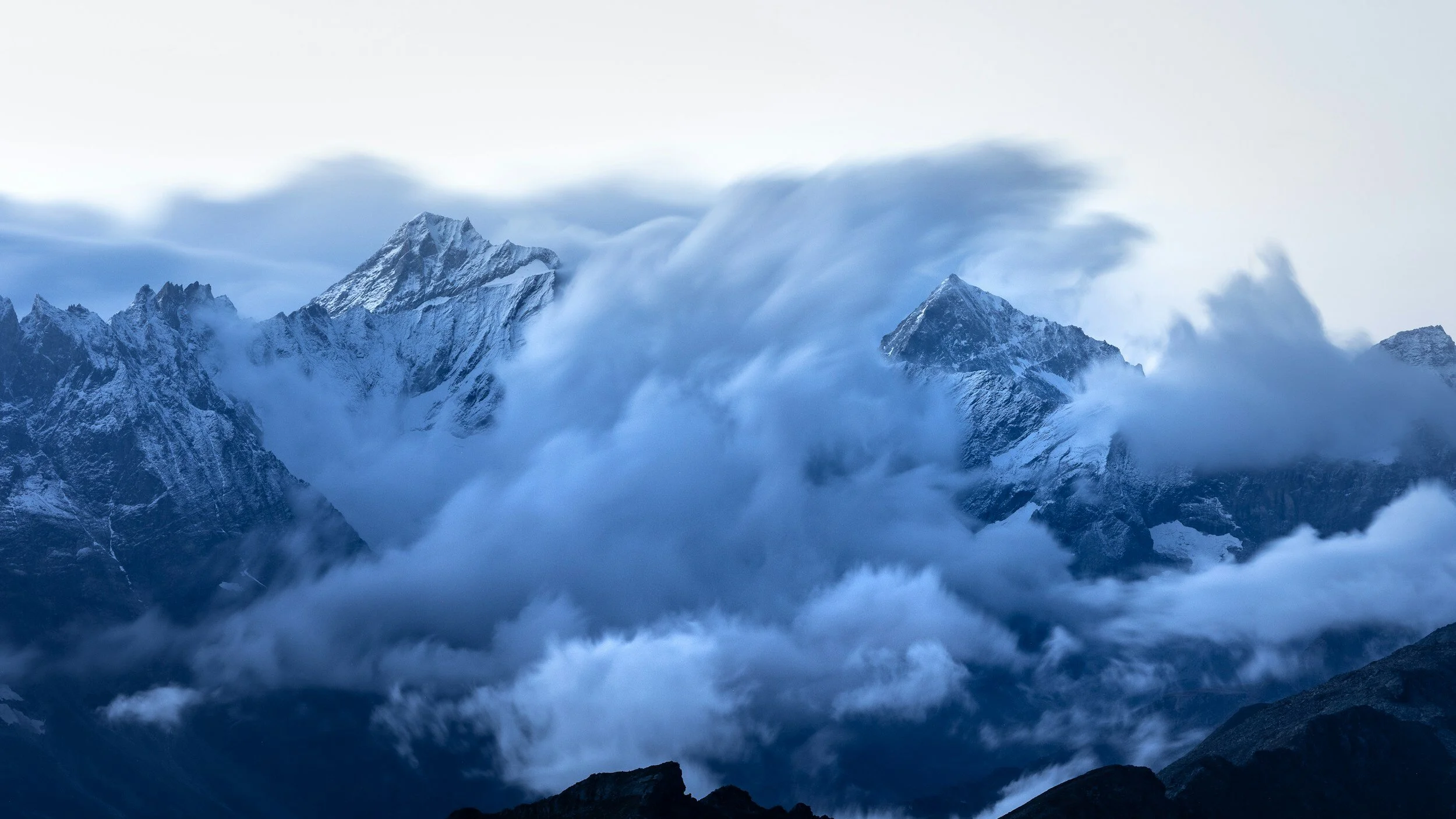 Snow-capped mountain peaks with clouds surrounding them in a rugged mountain landscape.