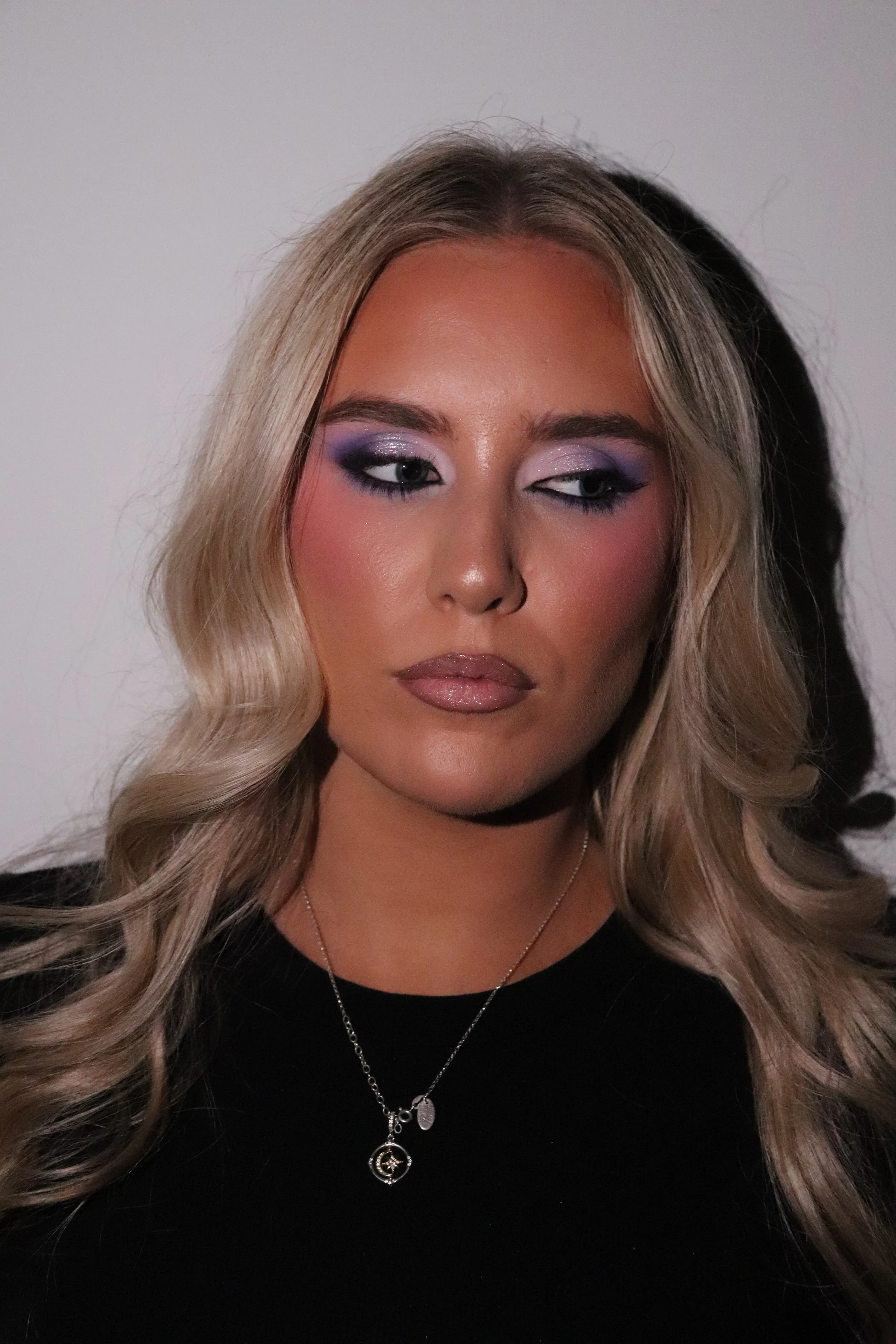 A woman with long, wavy blonde hair, wearing makeup with purple eyeshadow and nude lipstick, dressed in a black top, wearing layered necklaces, standing against a plain wall.