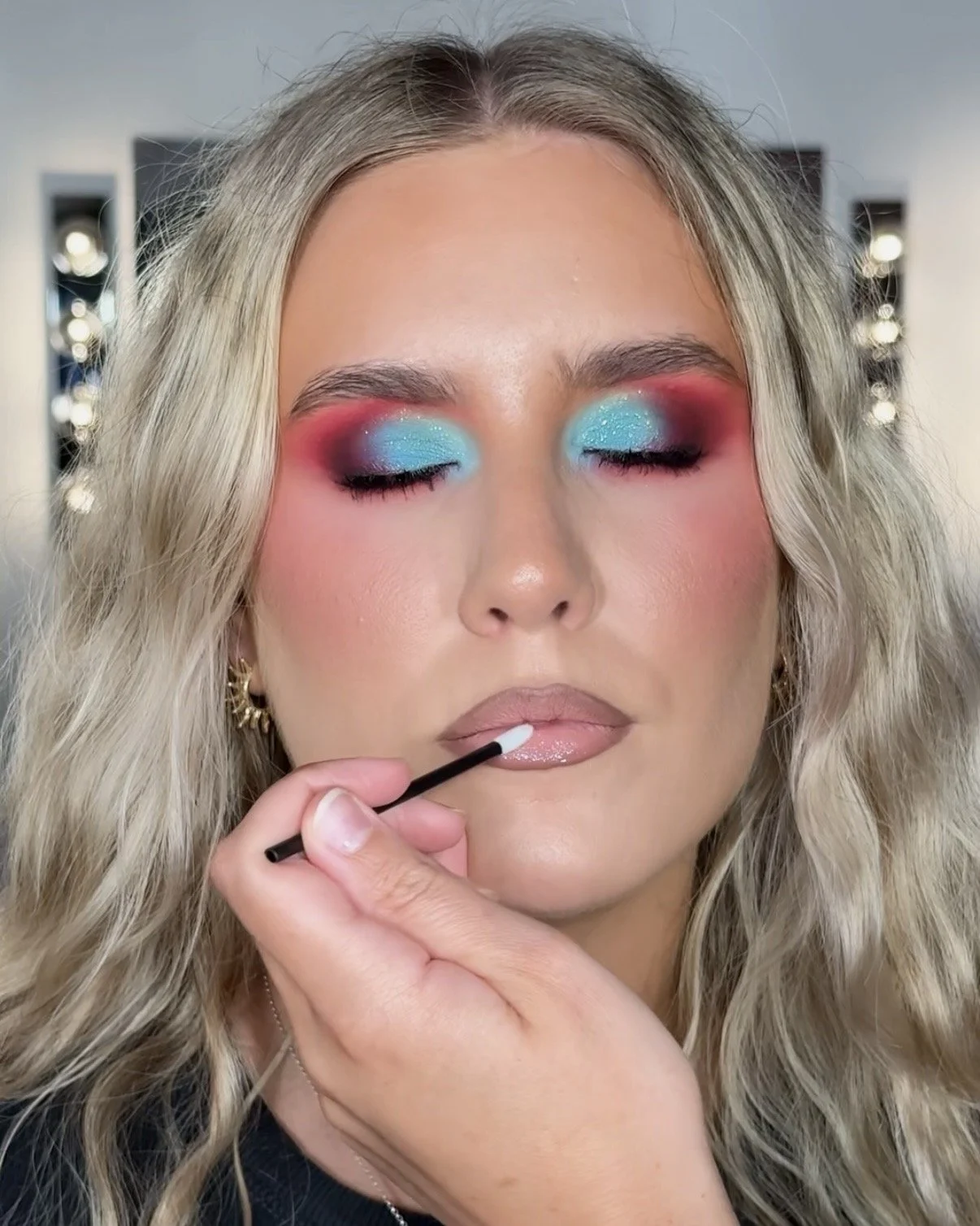 A woman with wavy blonde hair is applying lip gloss with a brush, her eyes closed, wearing vibrant eyeshadow in blue and pink shades, and gold earrings.