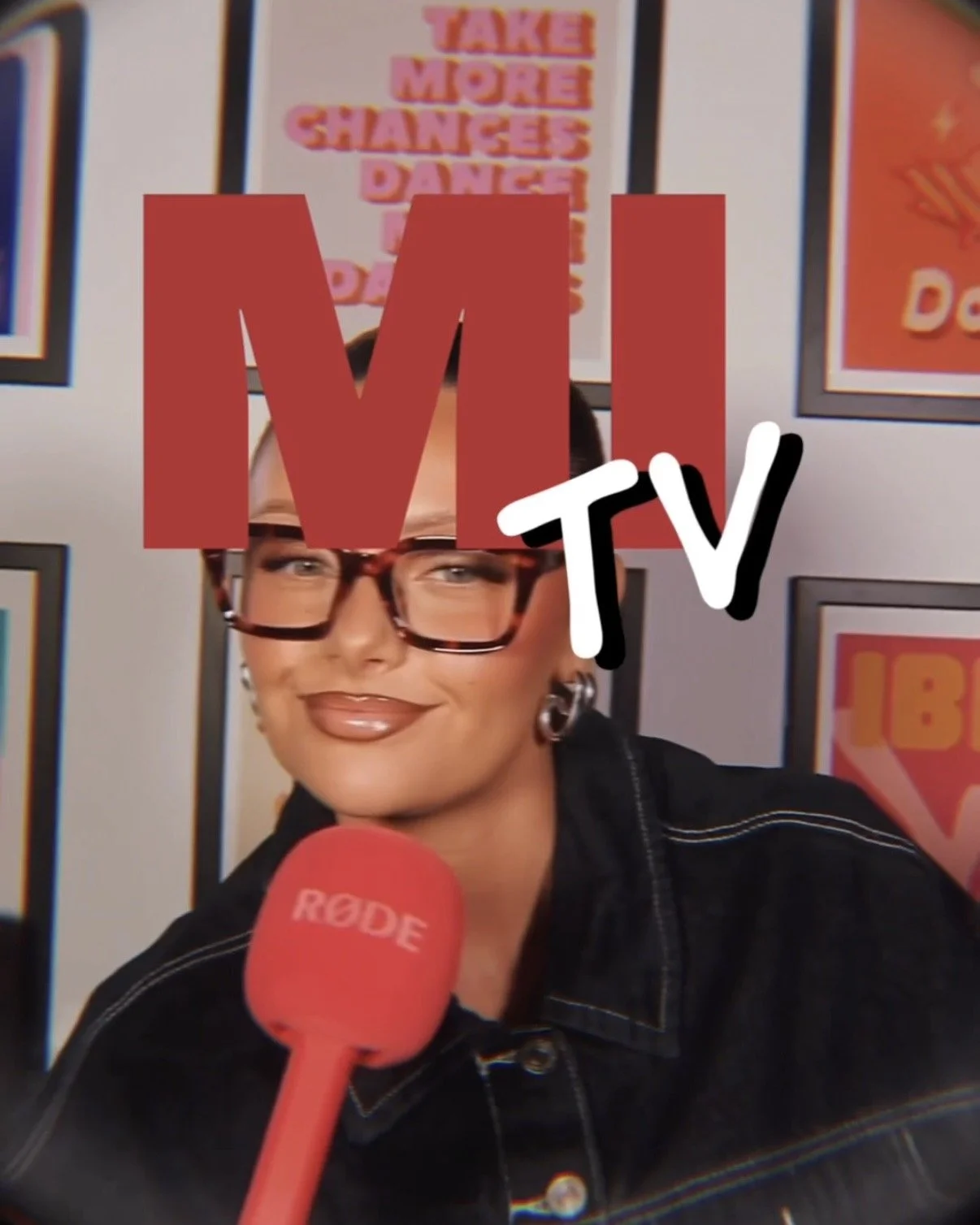 A woman with short dark hair, glasses, and earrings, speaking into a red microphone with the RØDE logo, in front of framed posters. The picture has a large red overlay of the letters "MI" with a white "TV" on top, partially covering her face.
