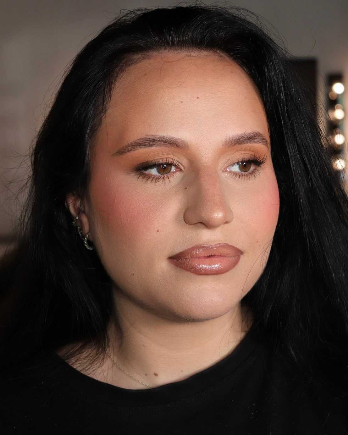 Close-up of a woman with long black hair and brown eyes, wearing makeup including eyeshadow, eyeliner, and glossy lips, with a black shirt and multiple earrings.
