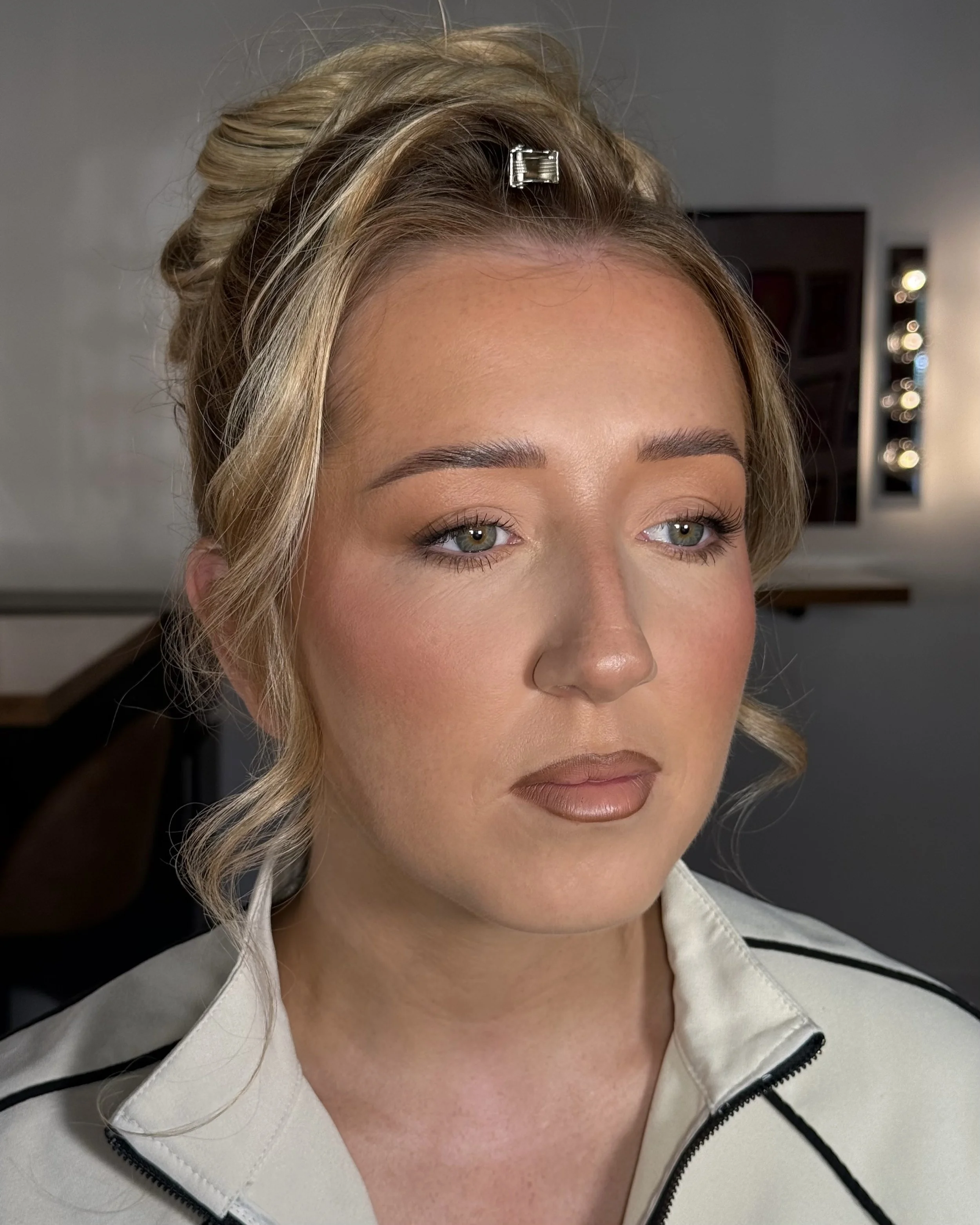 Close-up of a woman's face with blonde hair styled in loose waves, held back by a small silver hair clip. She has light skin, blue eyes, and subtle makeup. She's wearing a light-colored jacket with black accents.
