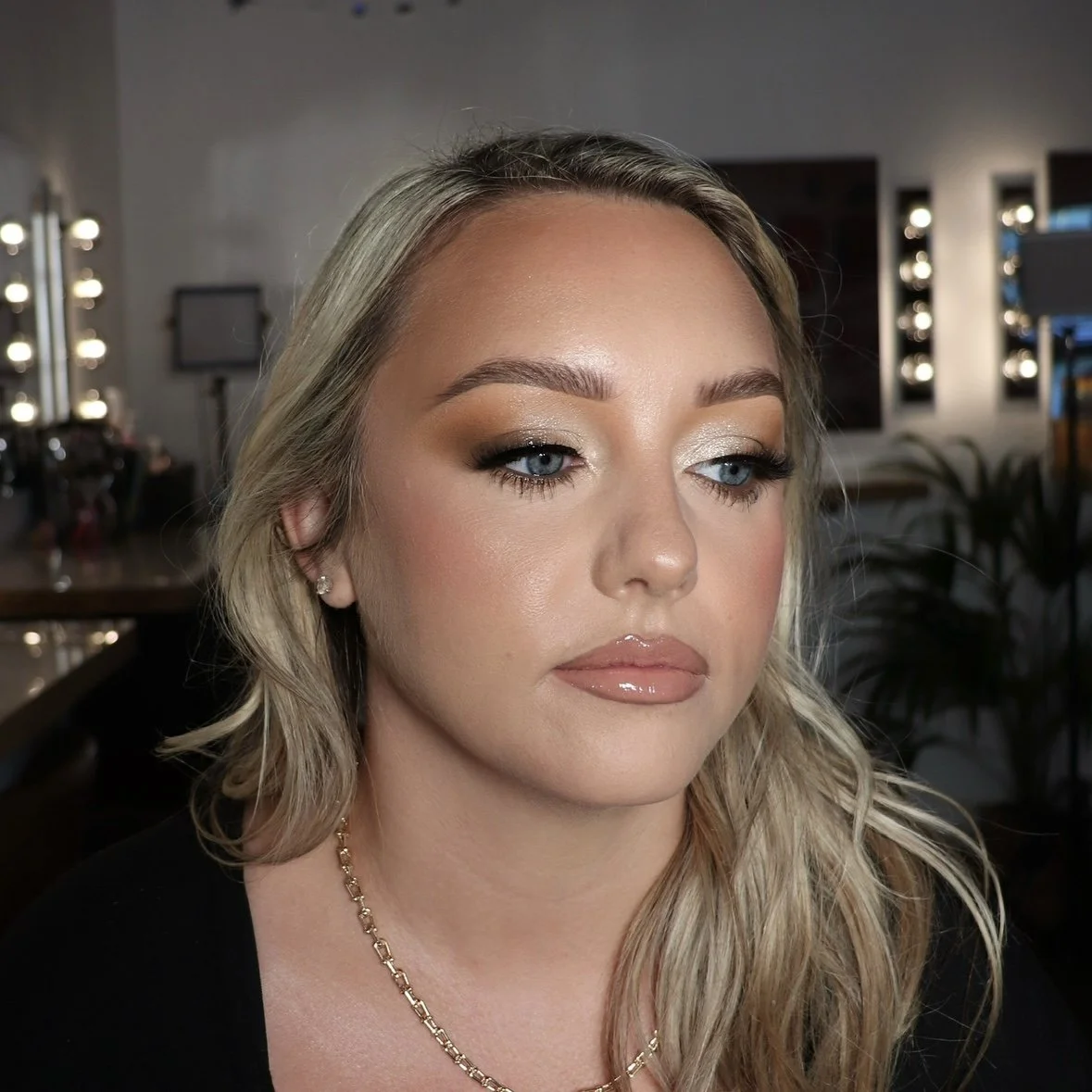 Close-up of a woman with blonde hair and makeup in a professional setting, with vanity lights and studio equipment in the background.