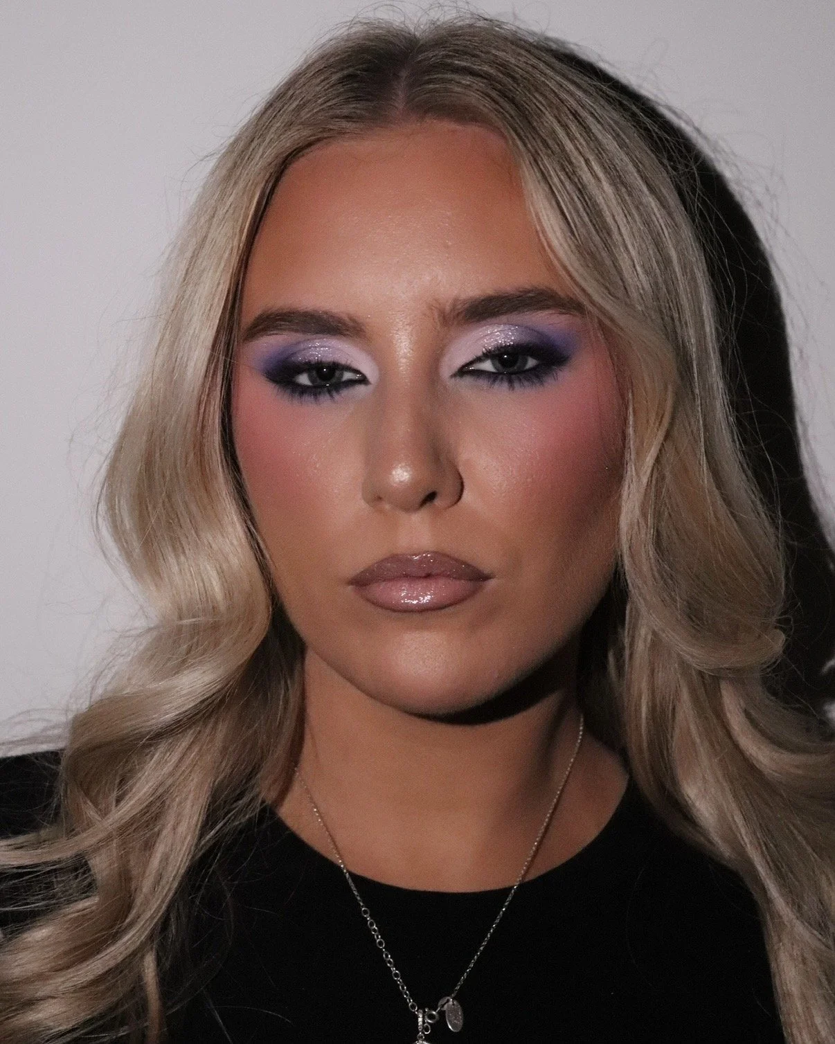 A woman with wavy blonde hair and makeup with purple eyeshadow and shiny lips.