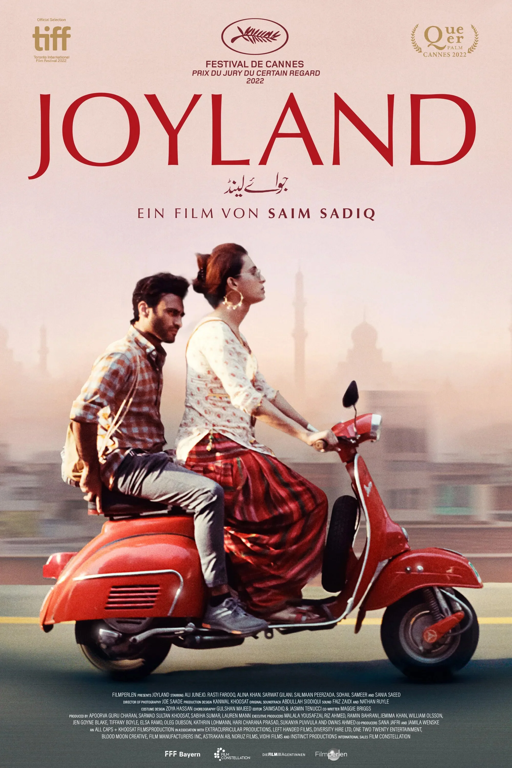A poster for the film 'Joyland' showing a man and woman riding a red scooter in front of a city skyline with a hazy pink sky. The woman is wearing glasses, earrings, a white top with a floral pattern, and a long red skirt. The man is wearing a checkered shirt and light-colored pants. The poster includes text in multiple languages, including English, German, and French, and logos for film festivals.