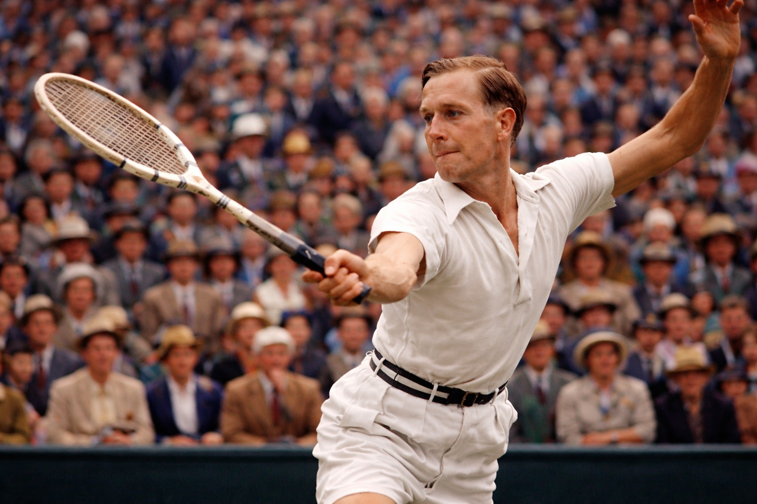 A male tennis player in a white outfit swinging his racket with an active crowd in the background.