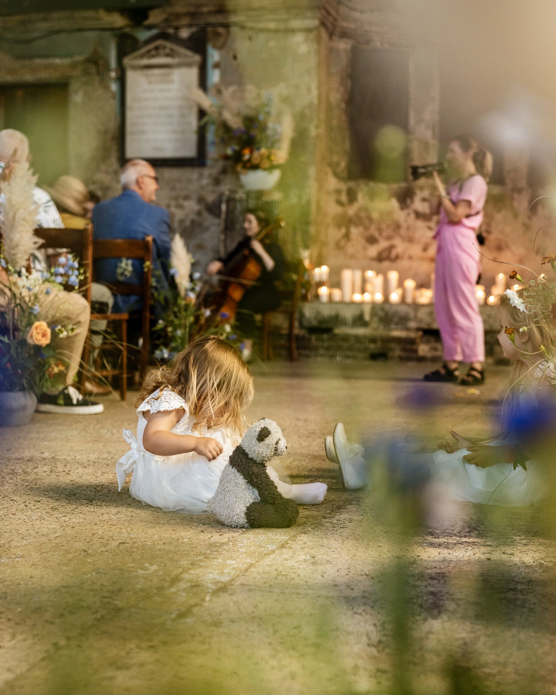 A young girl sits on a carpeted floor playing with a stuffed dog toy, with a group of people in the background attending a musical event in a rustic setting with candles and flowers.