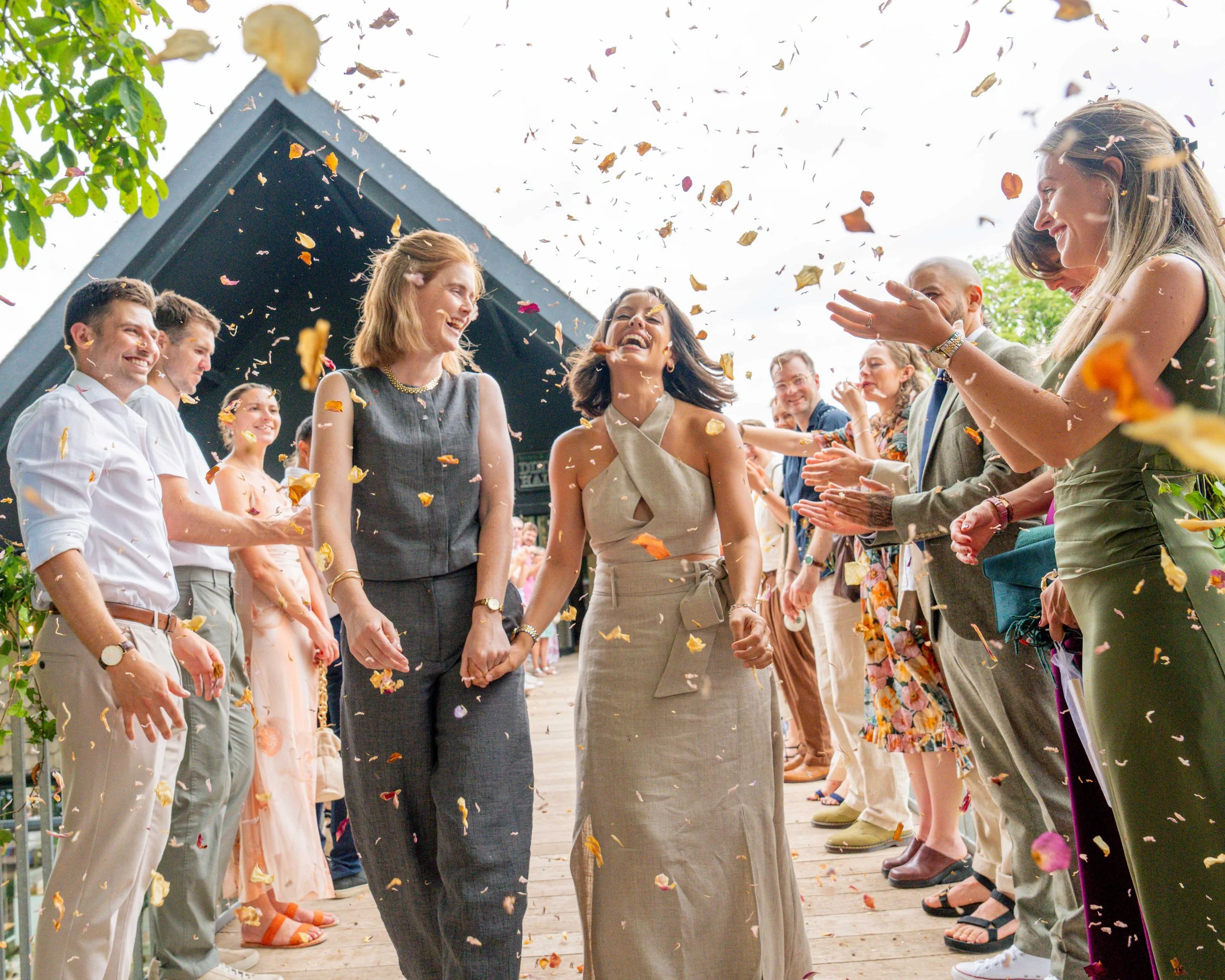 A group of people celebrating at a wedding, tossing flower petals while the bride and another woman hold hands and smile.
