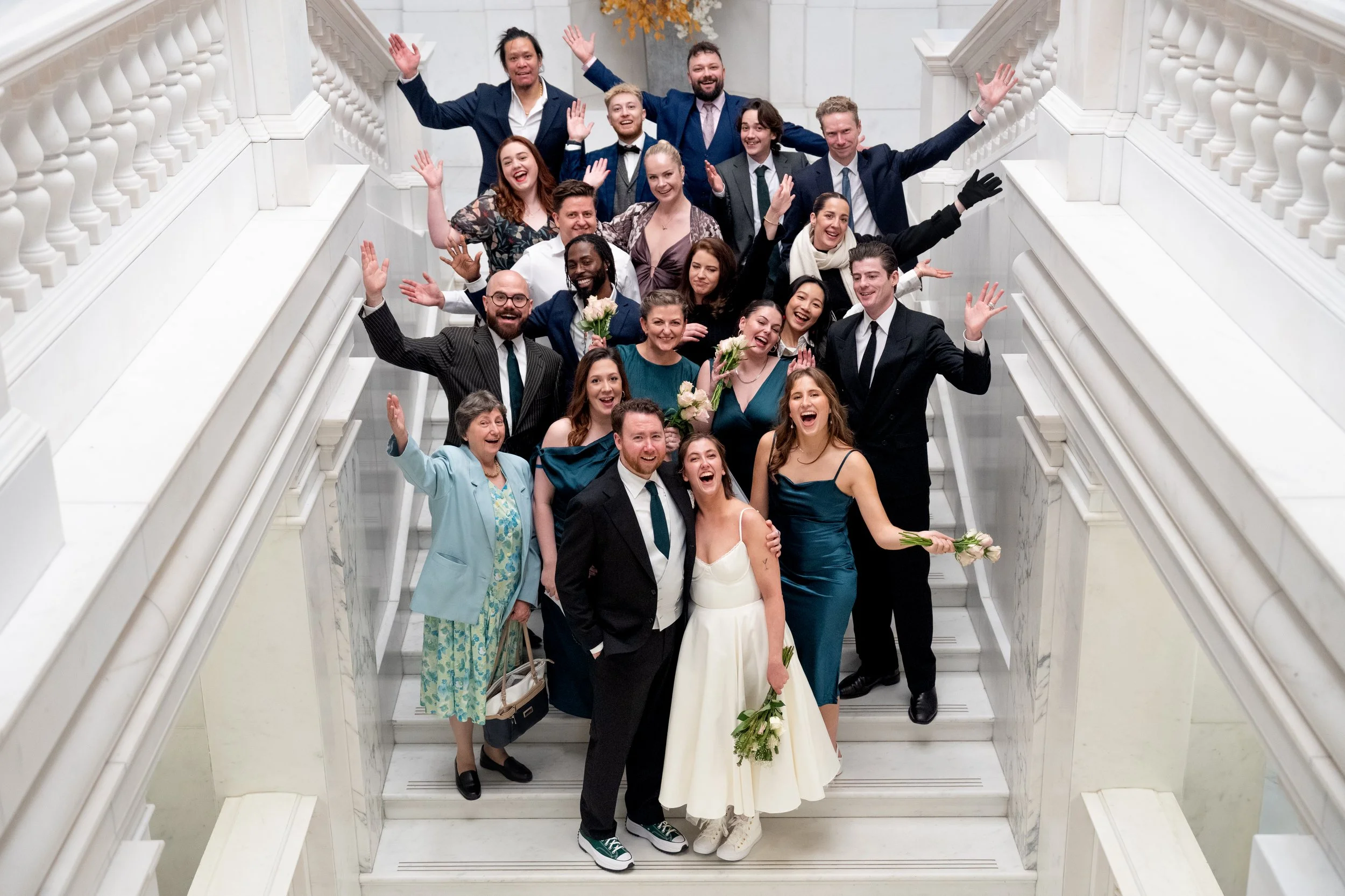 A large group of people dressed up in formal attire, standing on a marble staircase with white balustrades, celebrating and smiling at a wedding.