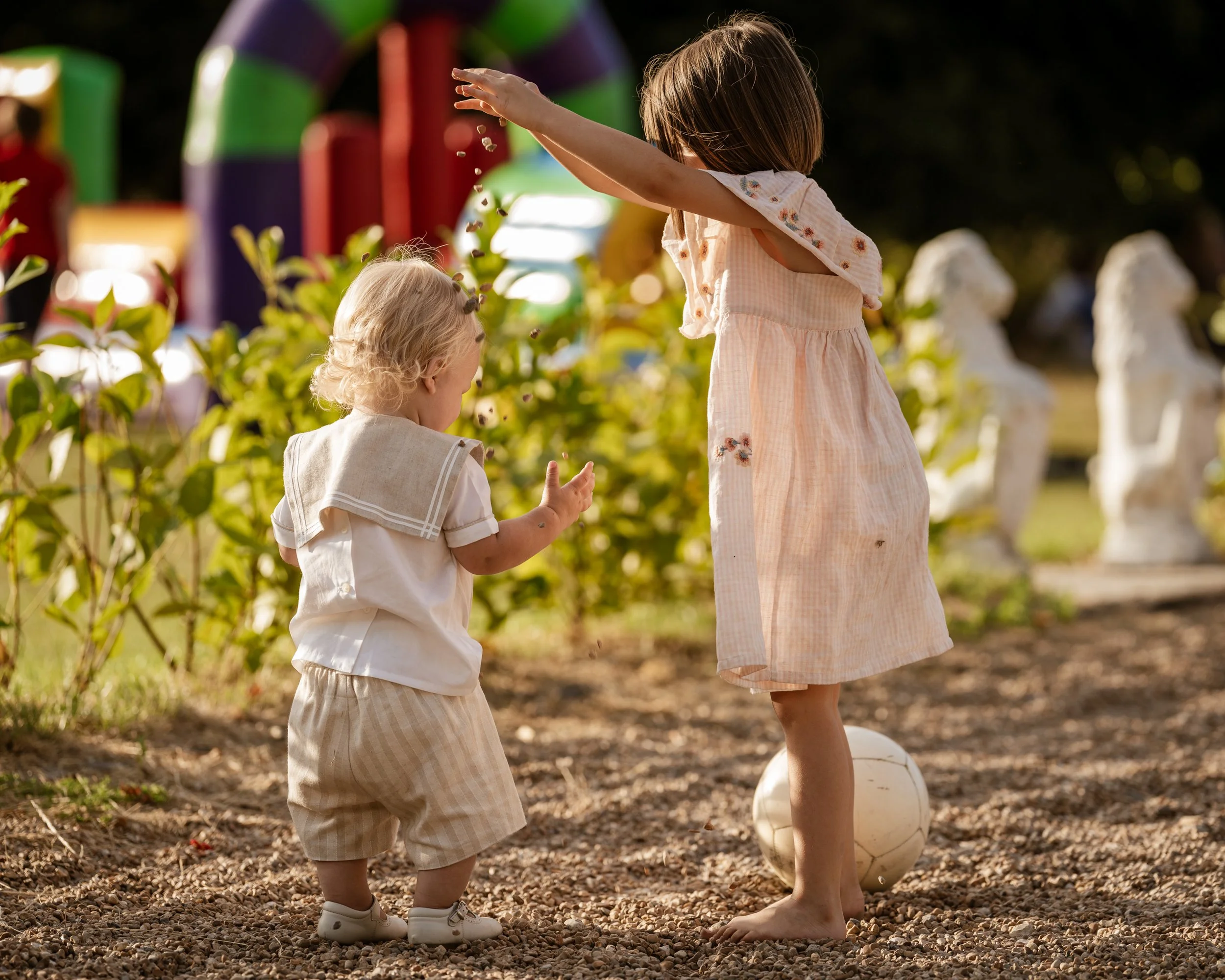 Two young girls playing with a ball outdoors in a garden or park setting during daytime, with colorful playground equipment in the background.