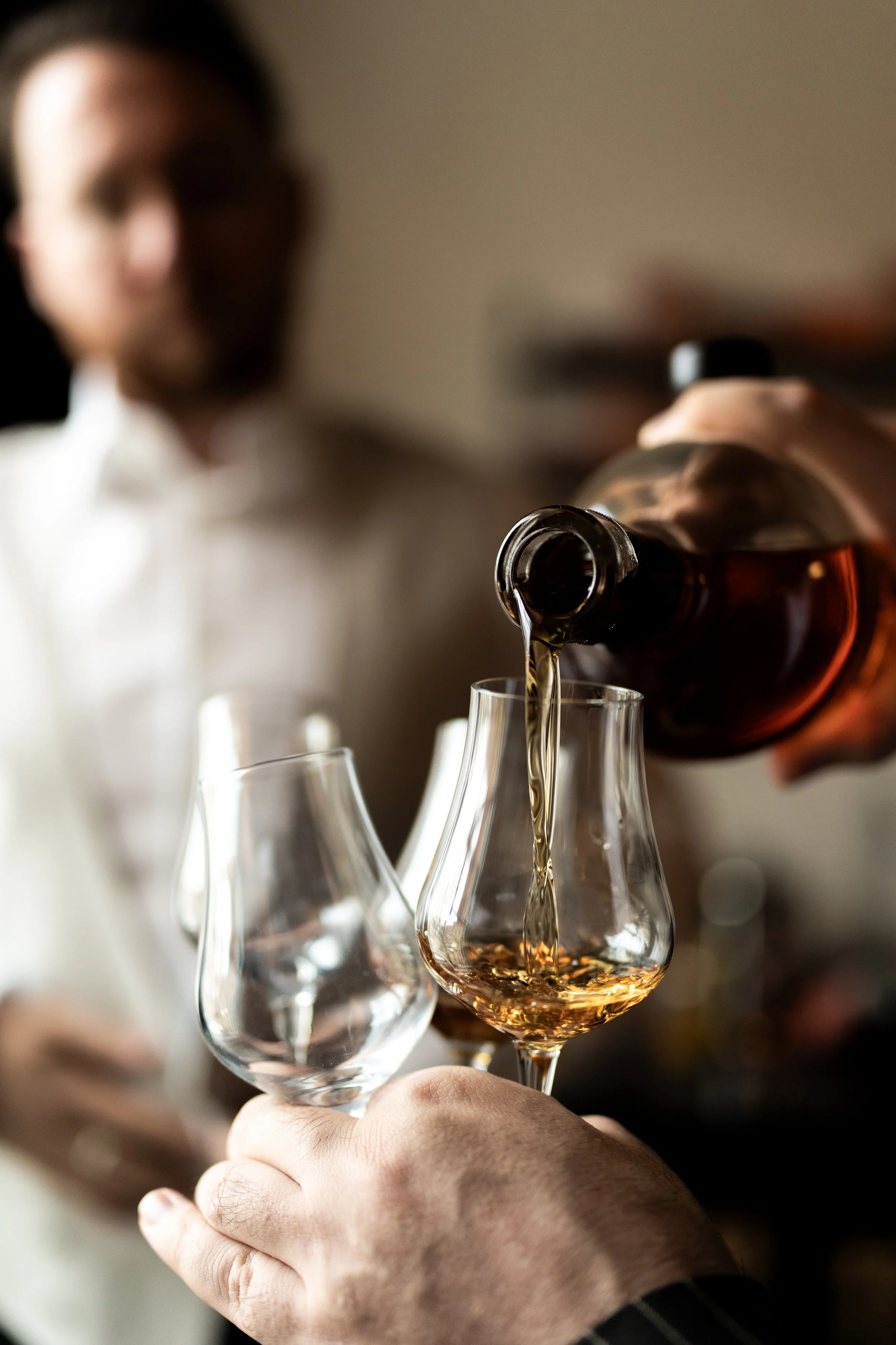 A groom, in a white shirt, and his best man pour whiskey into a glass held in a hand, with another empty glass nearby, in a dimly lit setting with a vintage feel.