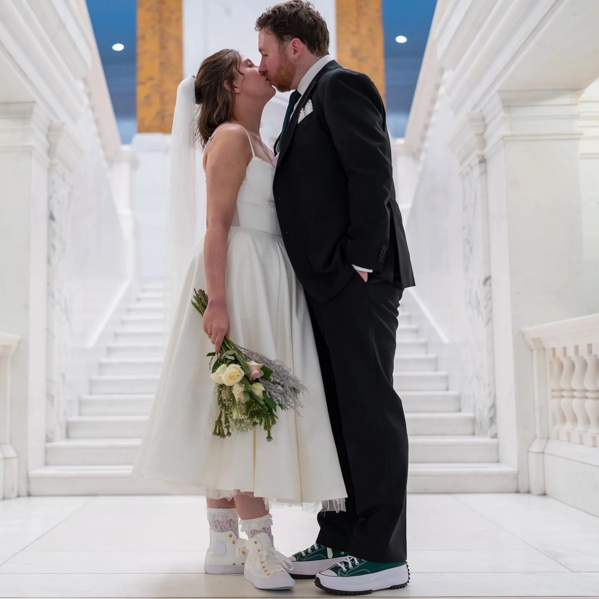 A bride and groom kissing on the steps of Camden Town hall. The bride is wearing trainers and holding a bouquet of yellow and white flowers