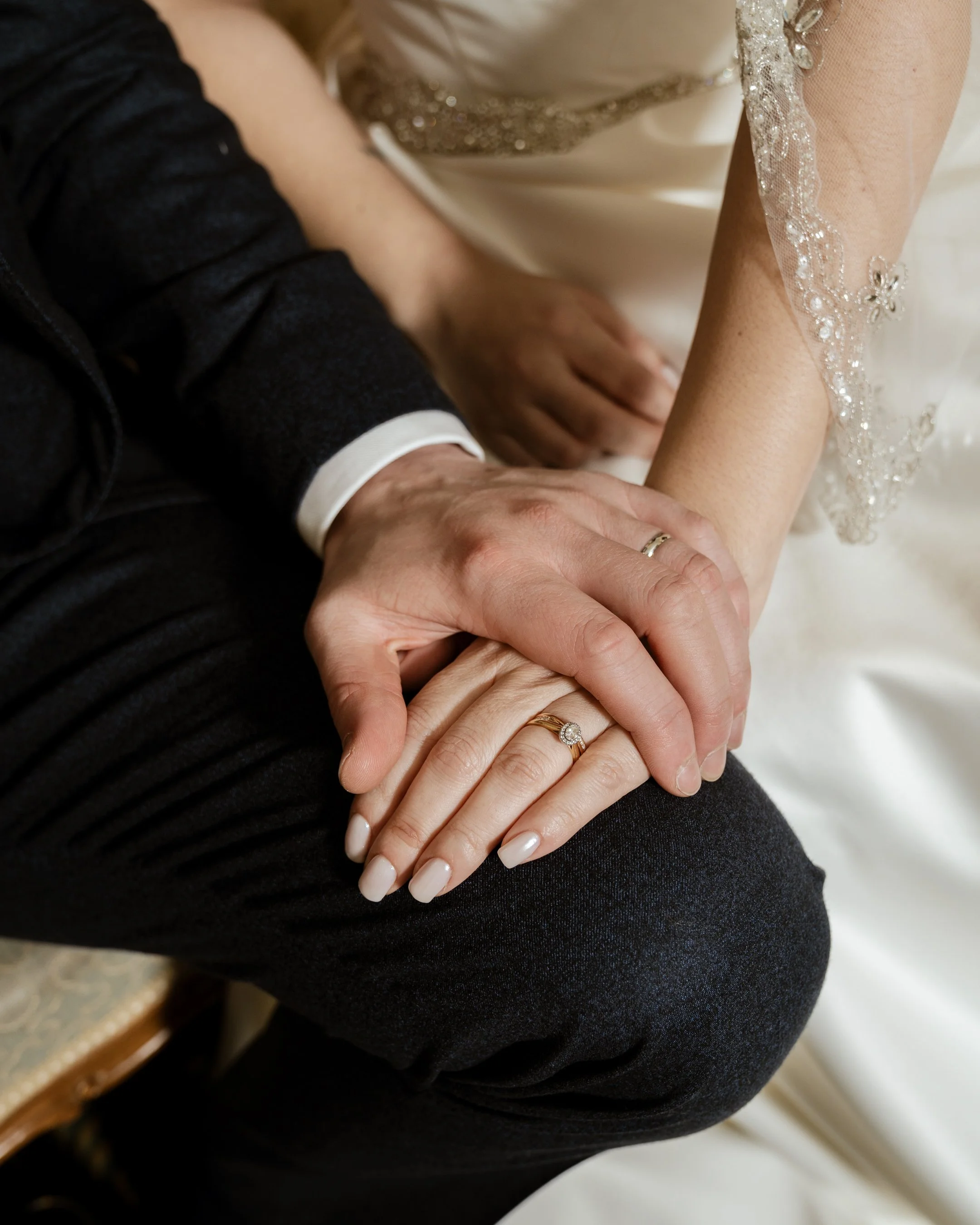 Close-up of a couple's hands with wedding rings, resting on a person's knee. The woman has a diamond engagement ring and the man has a wedding band. The woman's attire includes a white dress with beaded lace details, and the man's clothing is dark.