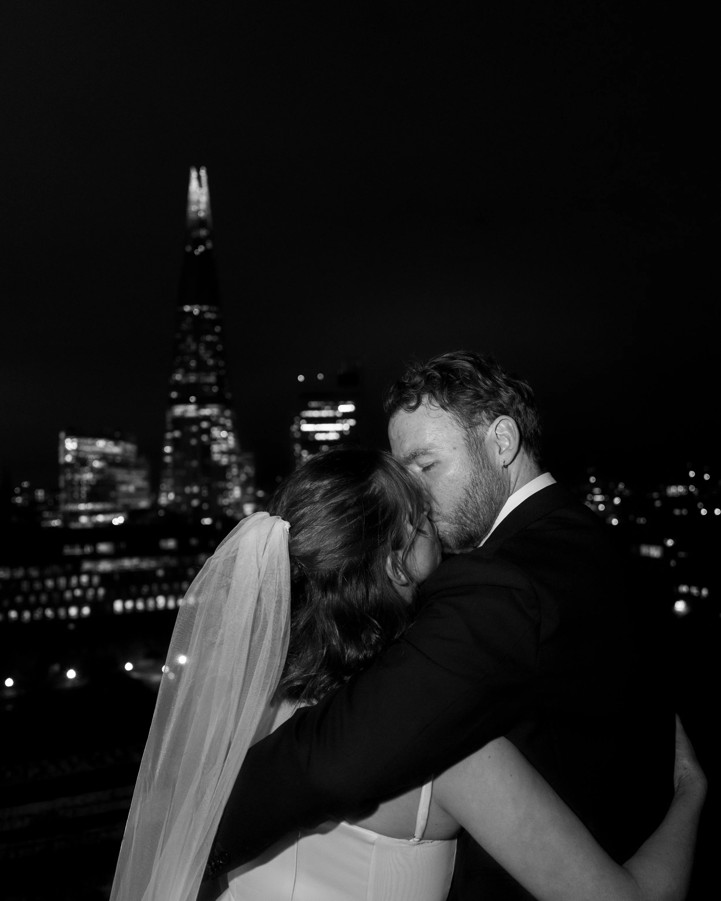 Black and white photo of a groom and bride kissing on a rooftop at night, with a city skyline and the Shard building illuminated in the background.