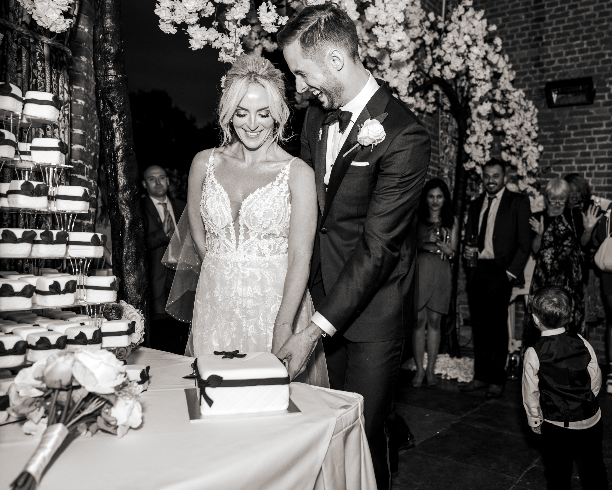 A bride and groom cut a wedding cake together at their London based wedding reception while guests watch and smile in the background.