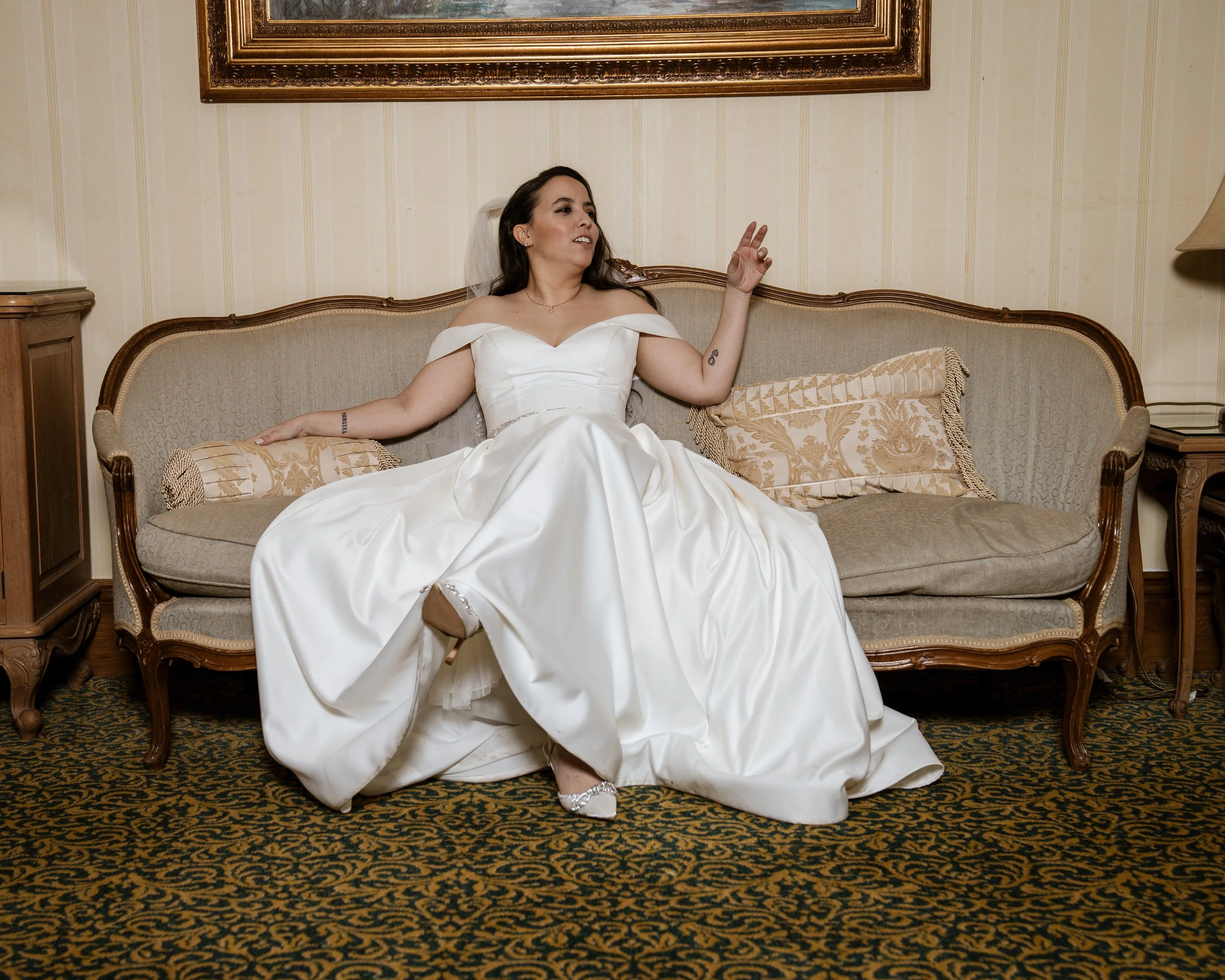 A woman in a white wedding dress sitting on a vintage sofa in a decorated room, with a framed painting above her, making a peace sign with her right hand.