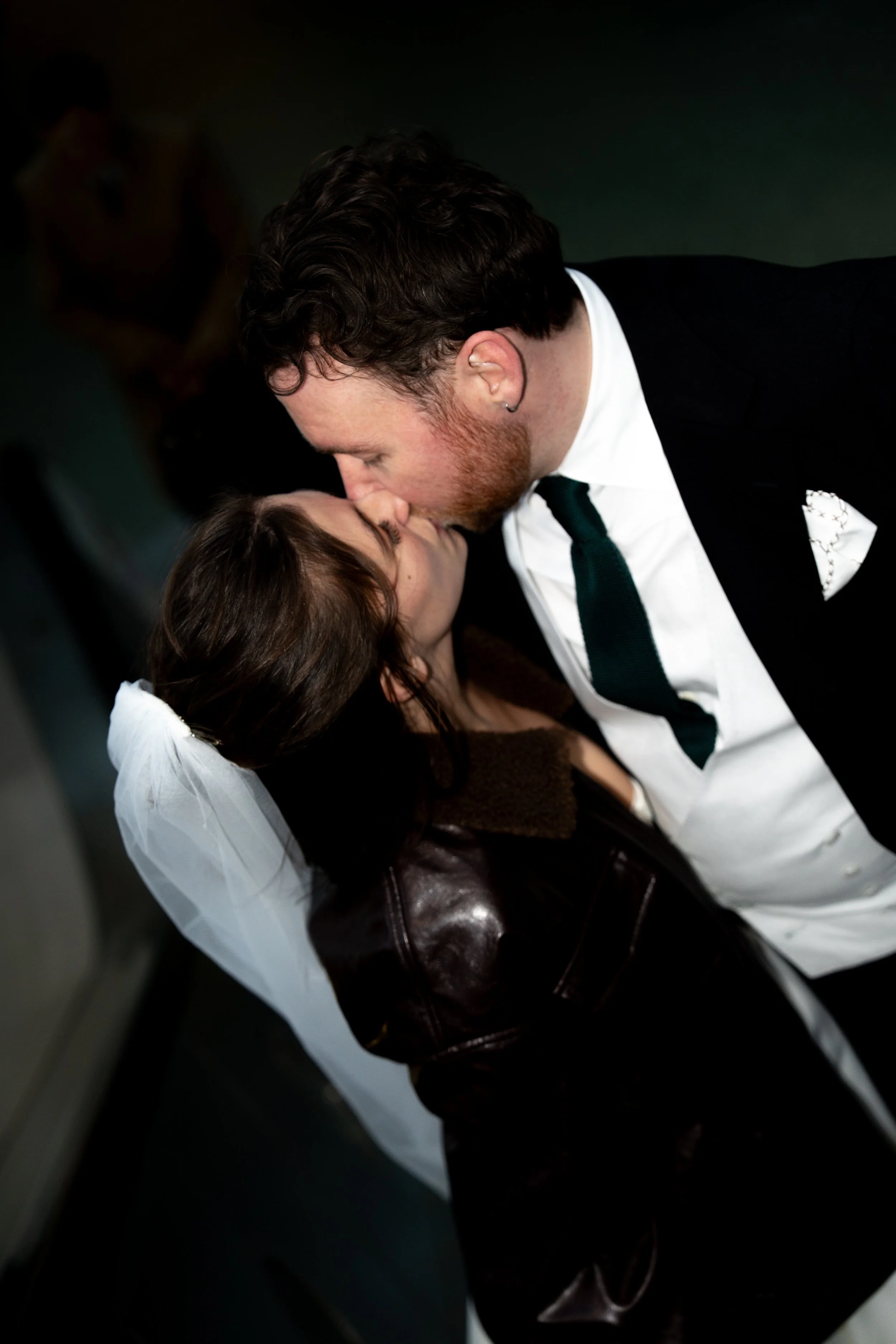 A couple sharing a kiss at a wedding, with the man wearing a suit and the woman in a leather jacket with a veil.
