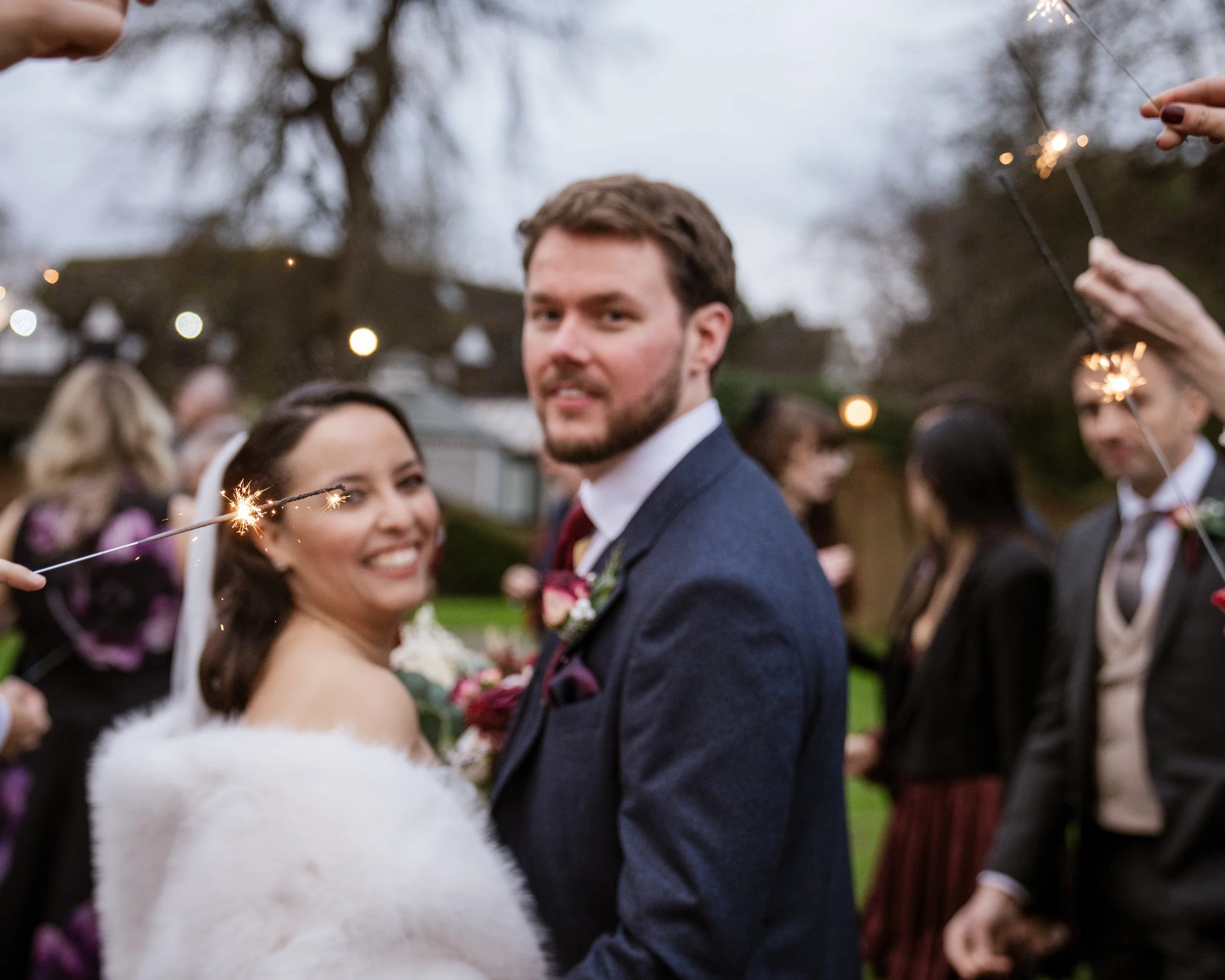 A smiling bride and groom at a wedding celebration, holding sparklers outside during twilight with guests in the background.