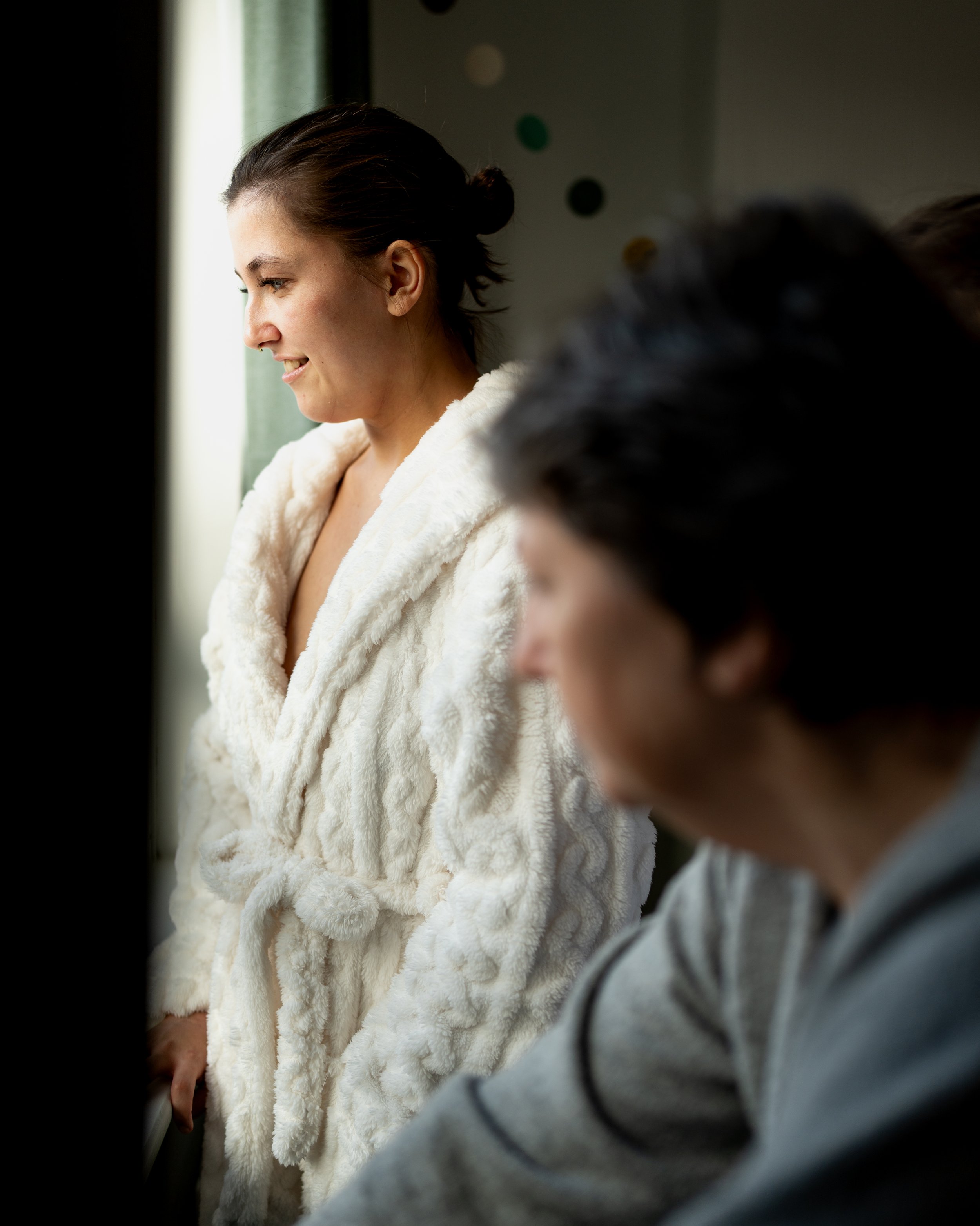 A woman in a white fuzzy coat looking out a window with a smile, with a man in a gray hoodie in the foreground, out of focus.