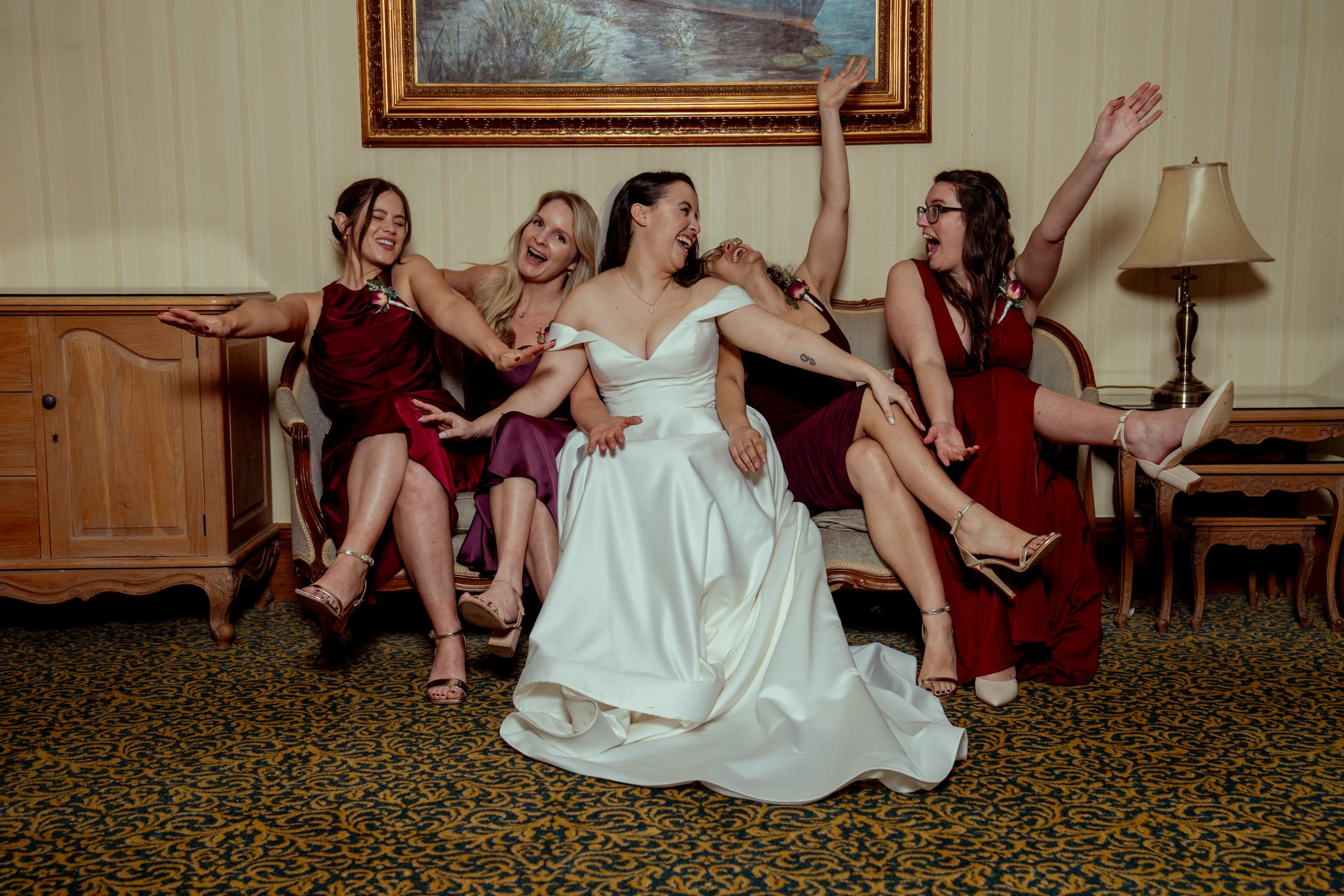 A bride and four bridesmaids laughing and celebrating her London based wedding with their arms raised and relaxed. The bride wears a white gown, while the bridesmaids wear maroon and purple dresses.
