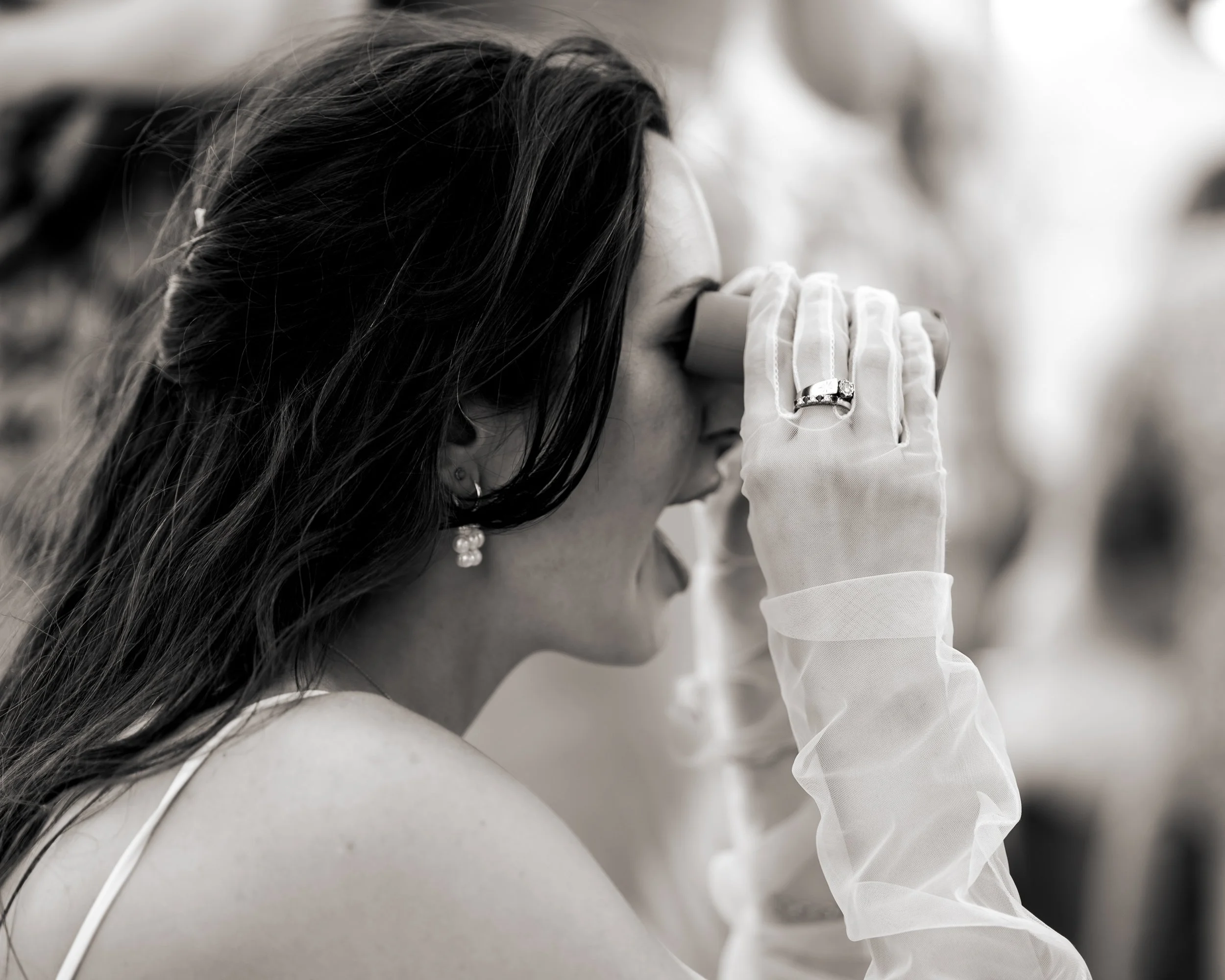 A woman in a wedding dress with long dark hair, wearing earrings and a wedding ring, holding her hand to her forehead with a confused or emotional expression.
