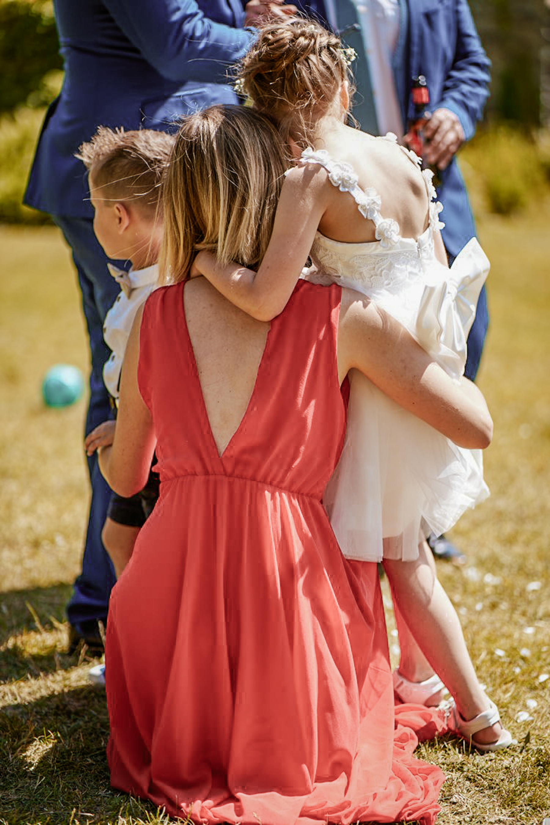 Group of children hugging and holding each other outdoors, with a woman in a red dress and a man in a blue suit in the background.
