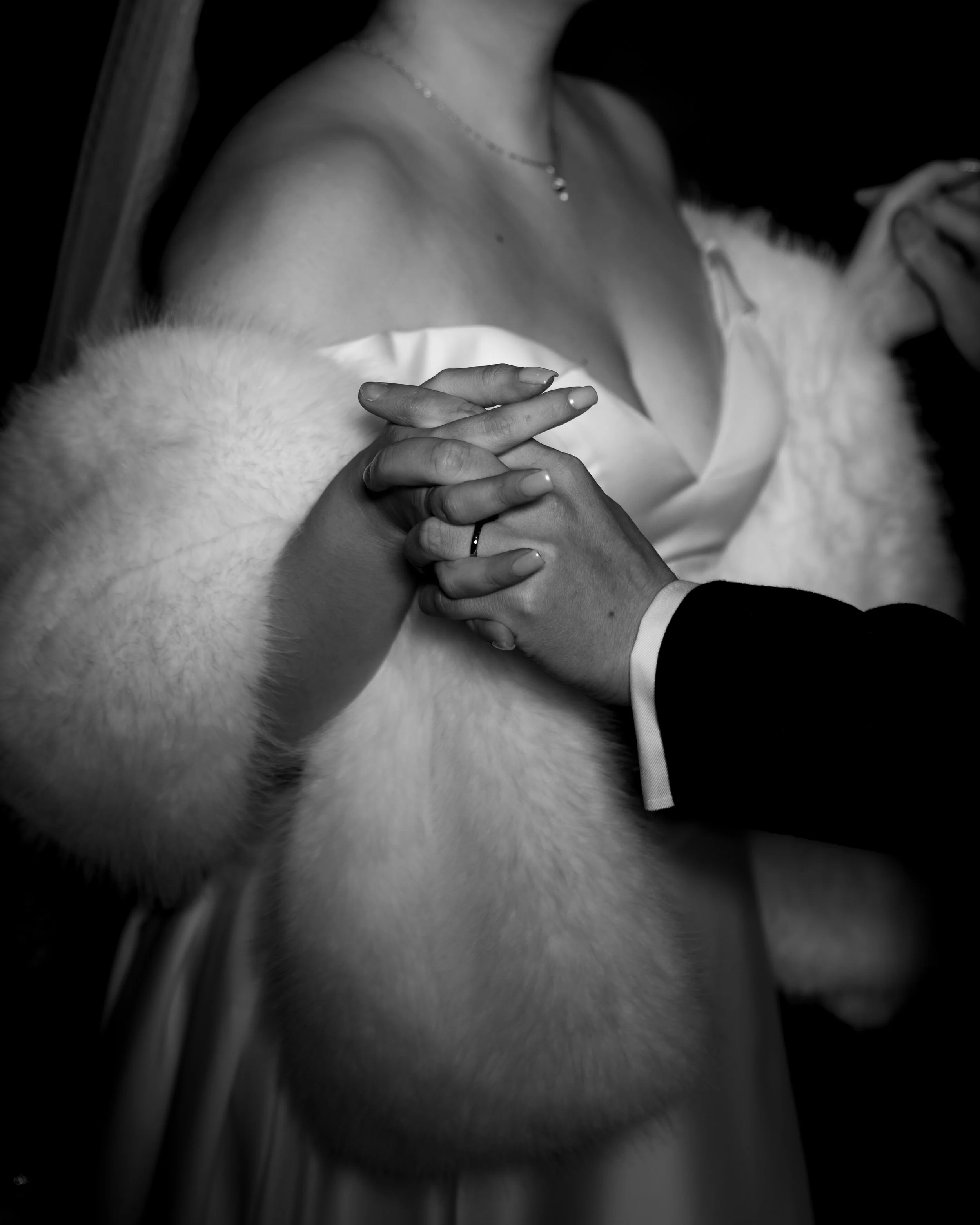 Close-up black and white photo of a couple holding hands during a wedding ceremony, with the woman wearing a white gown and fur shawl, and the man in a dark suit.