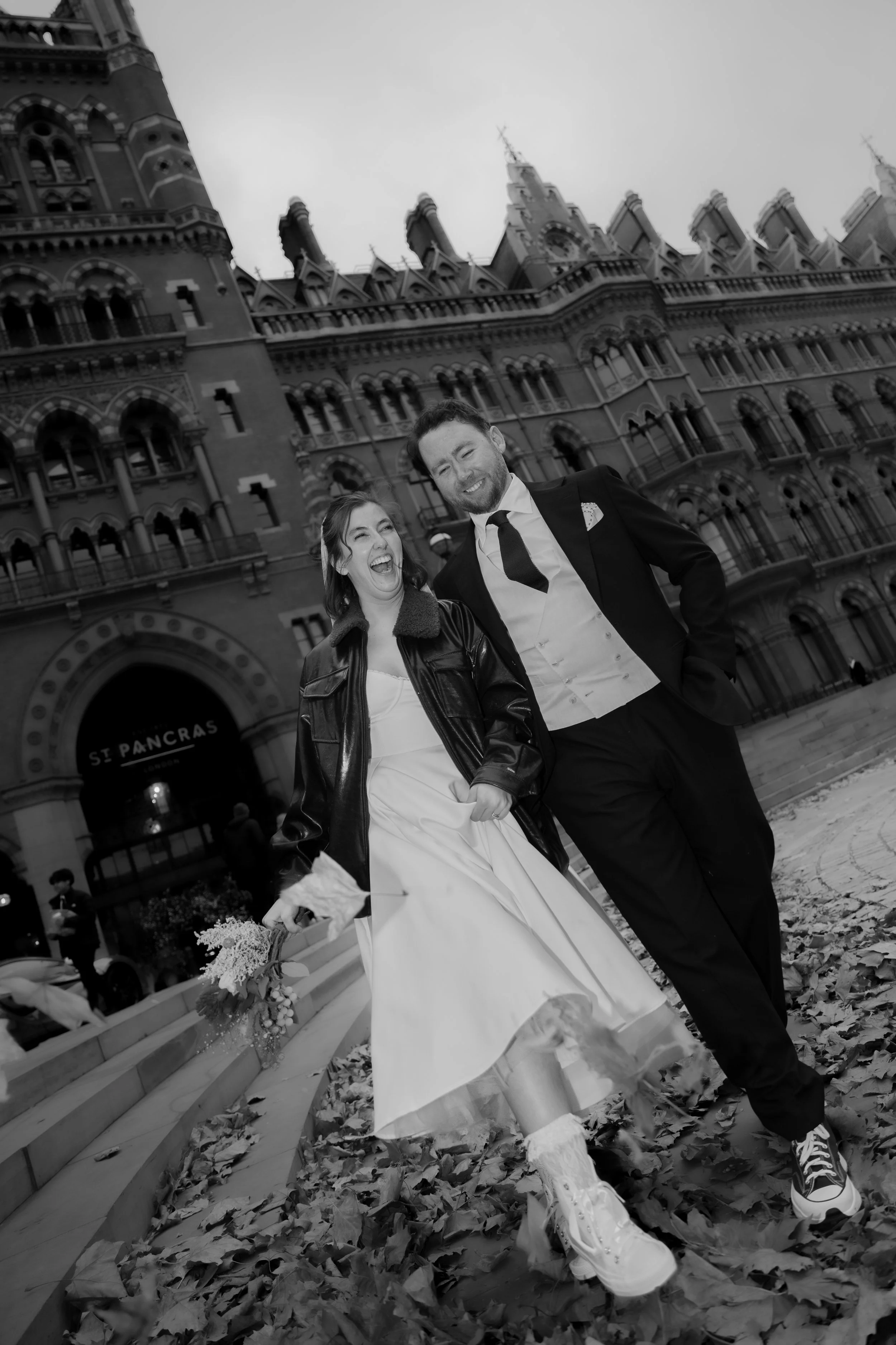 A smiling bride and groom couple dressed in their wedding attire walking in front of a historic building in Kings Cross, after their London wedding reception with fallen leaves on the ground, in black and white.