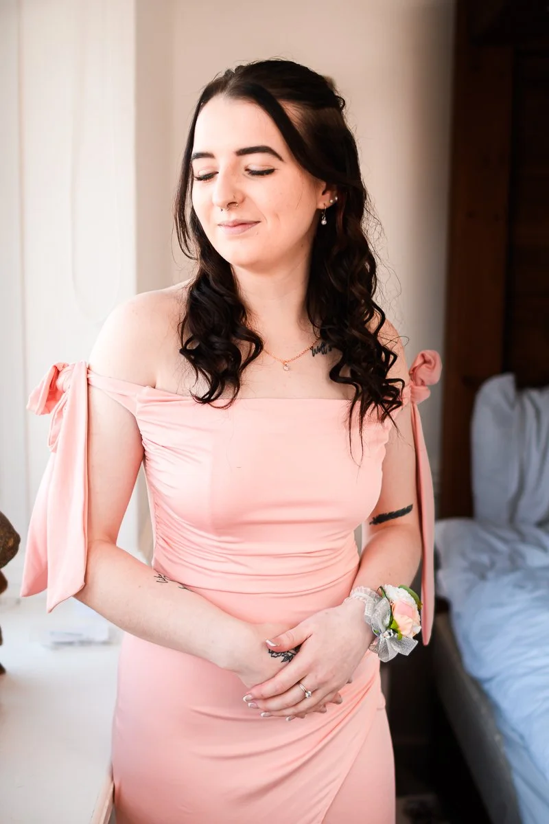 Woman in a peach off-shoulder dress with a corsage standing indoors, smiling with eyes closed and hands clasped.