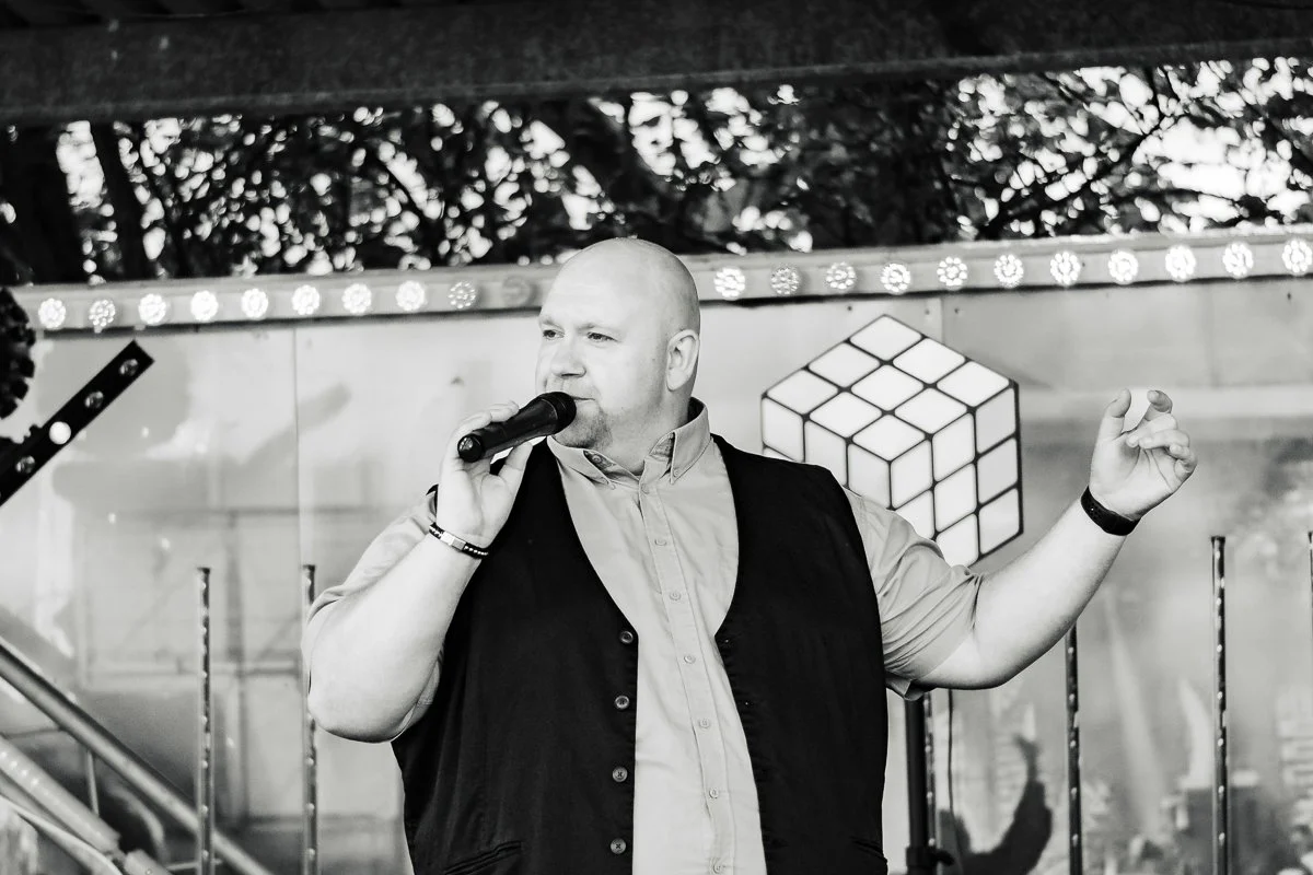 Man speaking into microphone on stage with Rubik's Cube backdrop.