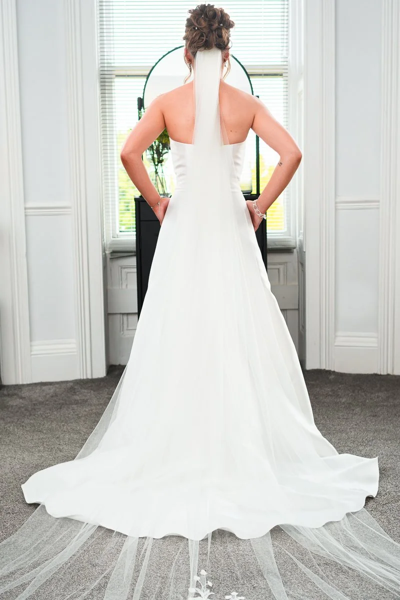 Bride in white wedding dress facing away with long veil, standing in a room with carpet and bright windows.