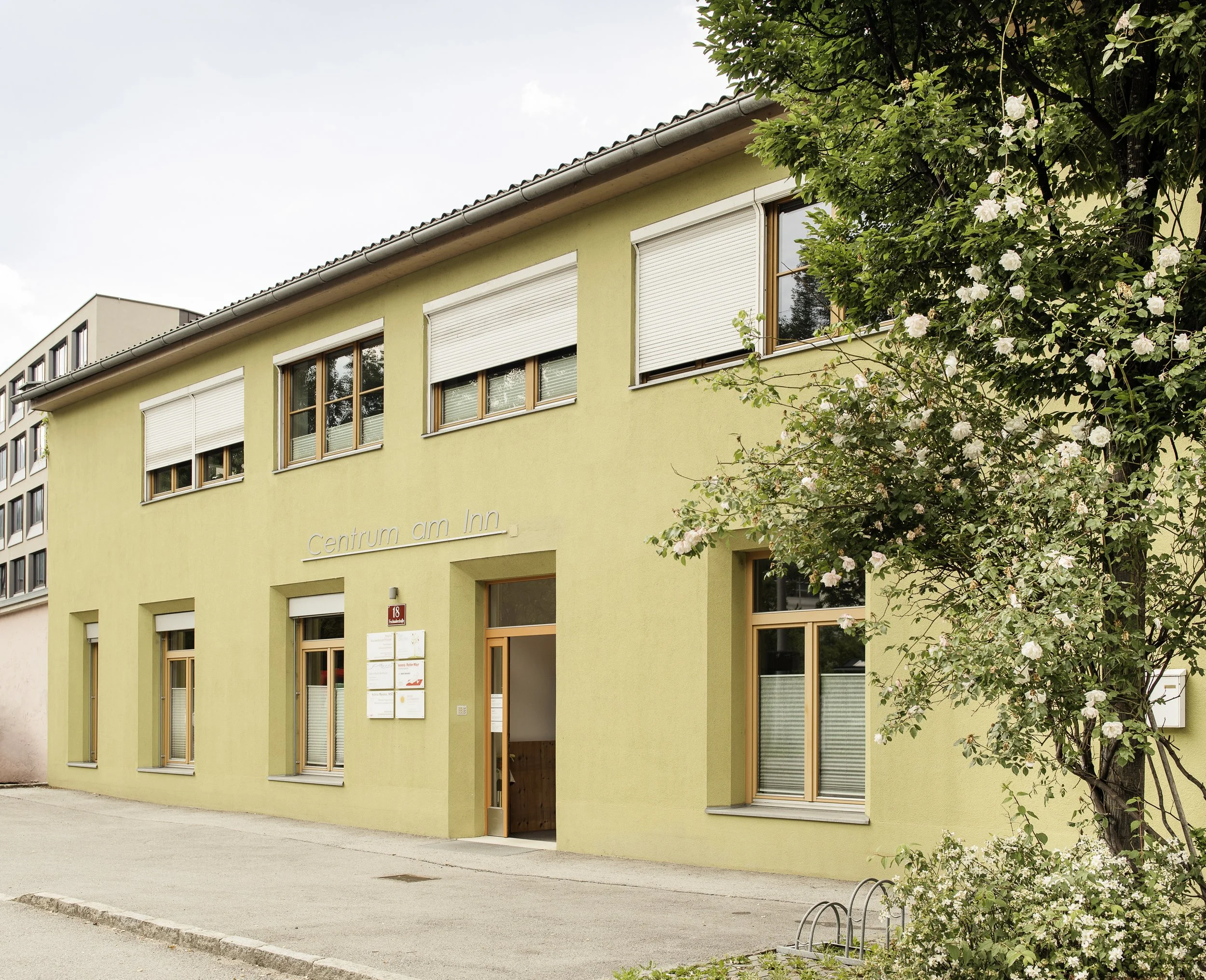 Ein zweistöckiges grünes Gebäude mit dem Namen 'Centrum am Inn' an einer Straßenfront, umgeben von Bäumen und einer Blumenhecke.