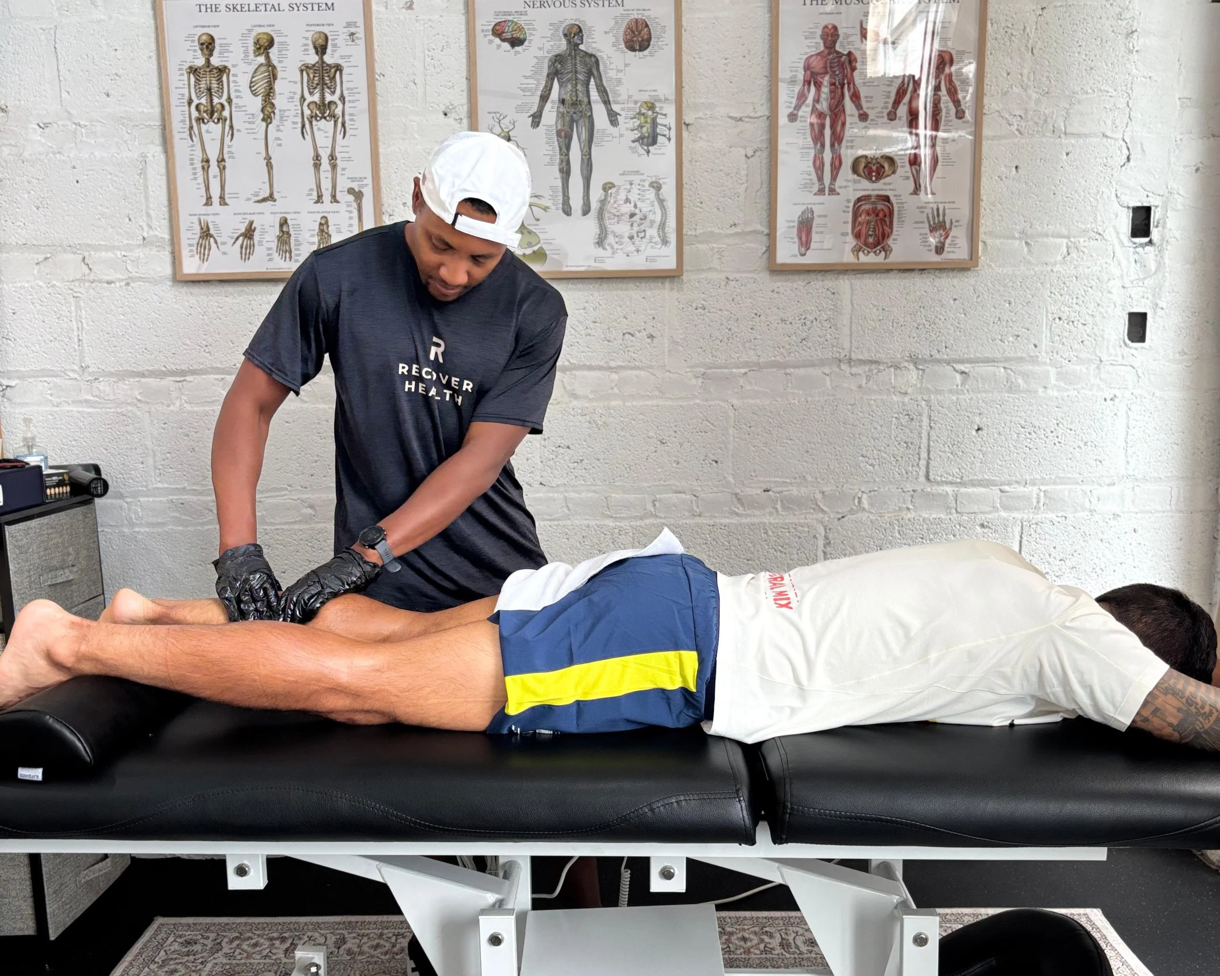 A person receiving a massage on their lower back and legs from a practitioner in a clinic with anatomical posters on the wall.