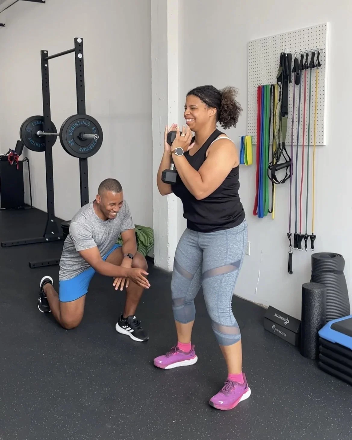 A woman holding a dumbbell and smiling at a man kneeling and laughing in a gym.