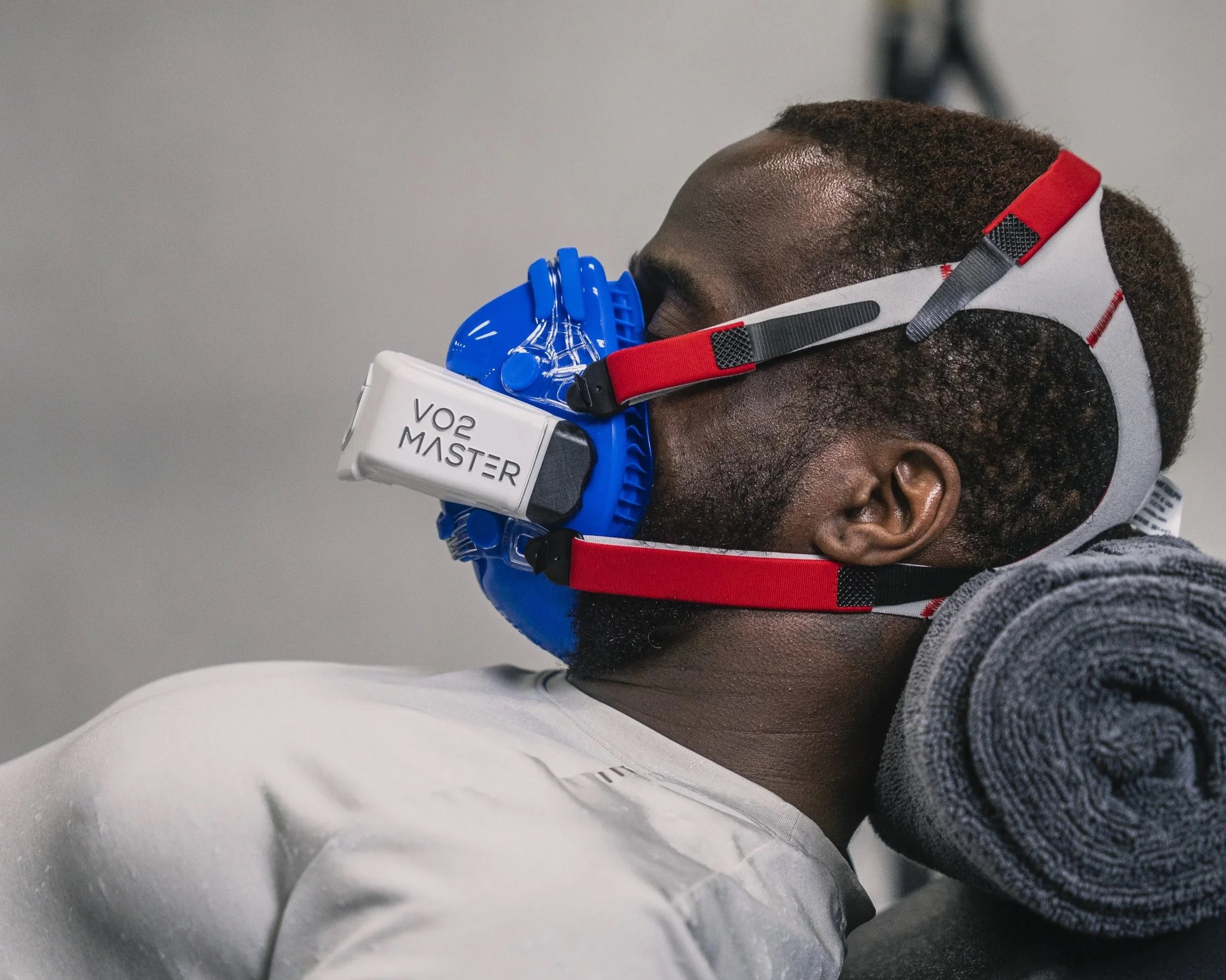 A man wearing a mask with a white mouthpiece labeled 'VO2 MASTER' and a head strap, resting with a gray towel rolled up against a neutral background.