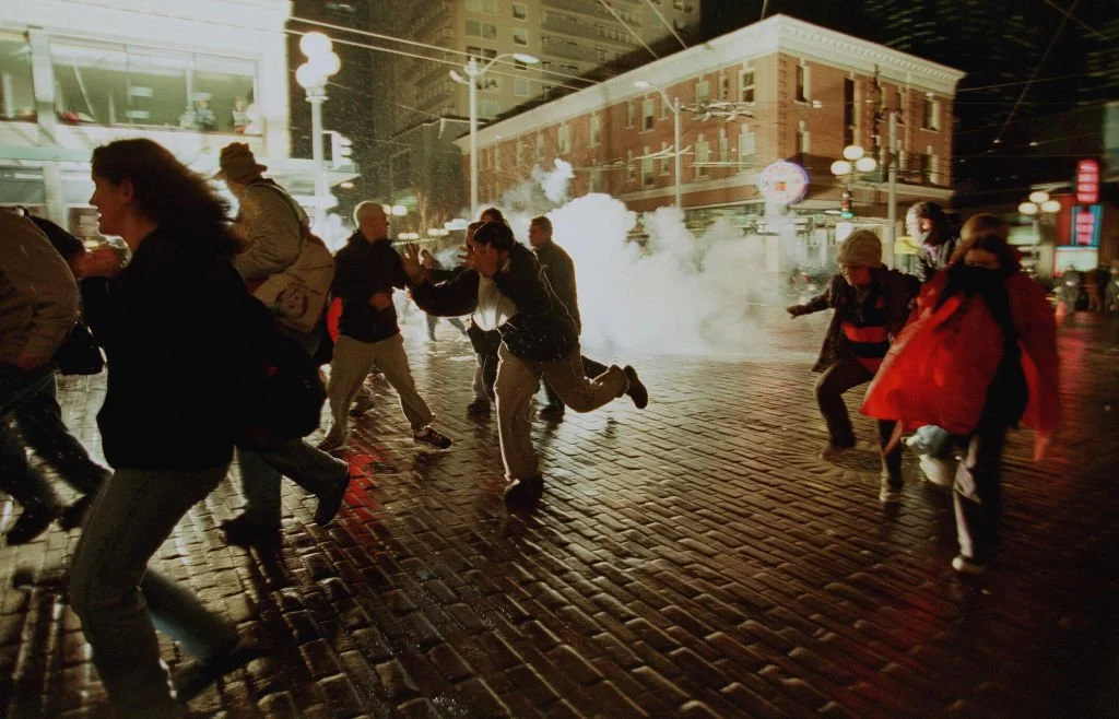 Menschen, die bei Nacht in einer Stadt auf nasser Kopfsteinpflasterstraße vor Tränengas fliehen.