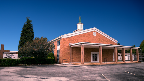Red brick church with a white steeple, surrounded by trees, under a clear blue sky.