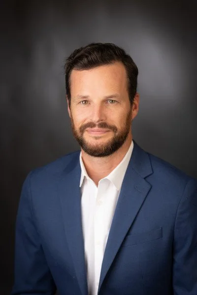 Headshot of a man with dark hair and beard wearing a blue suit and white shirt against a dark background.