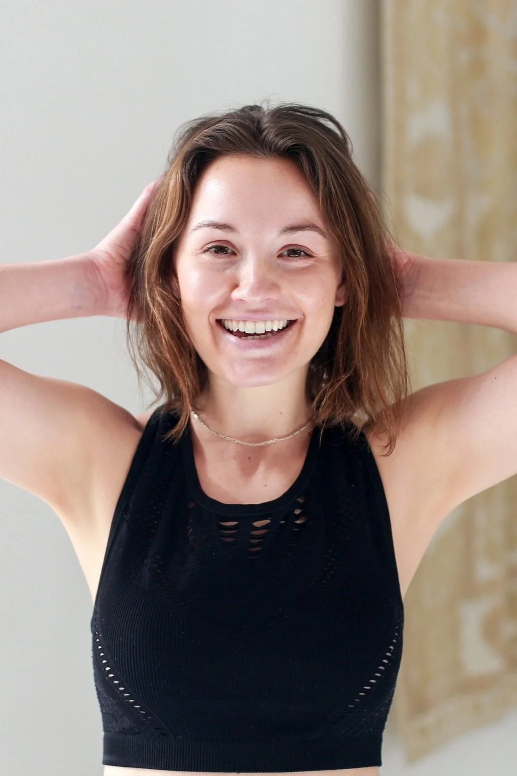 Young woman smiling, wearing a black sleeveless top, with hands behind her head.