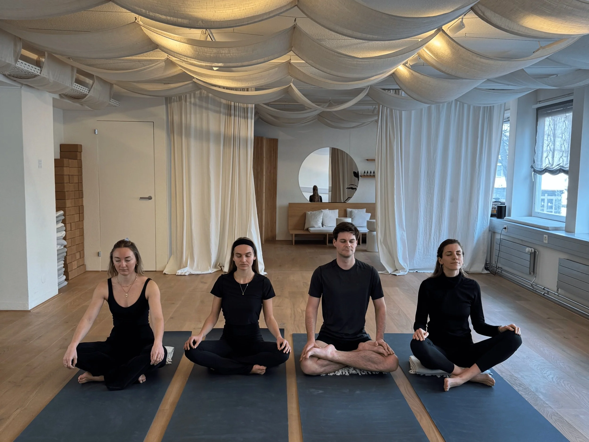 Four people practicing meditation in a peaceful, well-lit room with large windows, white curtains, and wooden flooring. They are seated on yoga mats in a cross-legged position with eyes closed.