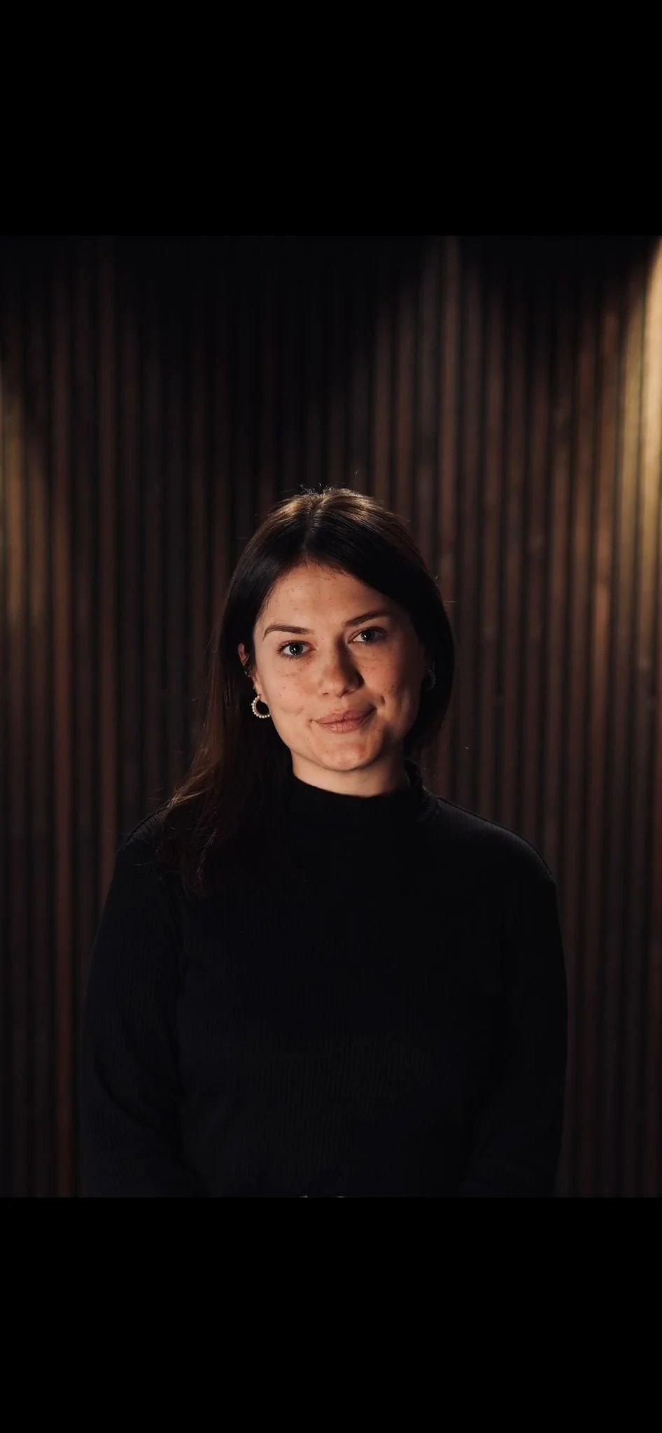 Portrait of a young woman with dark hair, wearing hoop earrings and a black turtleneck, standing in front of a wooden wall.