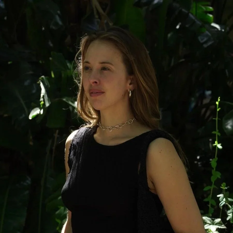 A woman with reddish-brown hair wearing a black sleeveless top and silver jewelry standing outdoors in front of lush green plants, with sunlight highlighting her face.