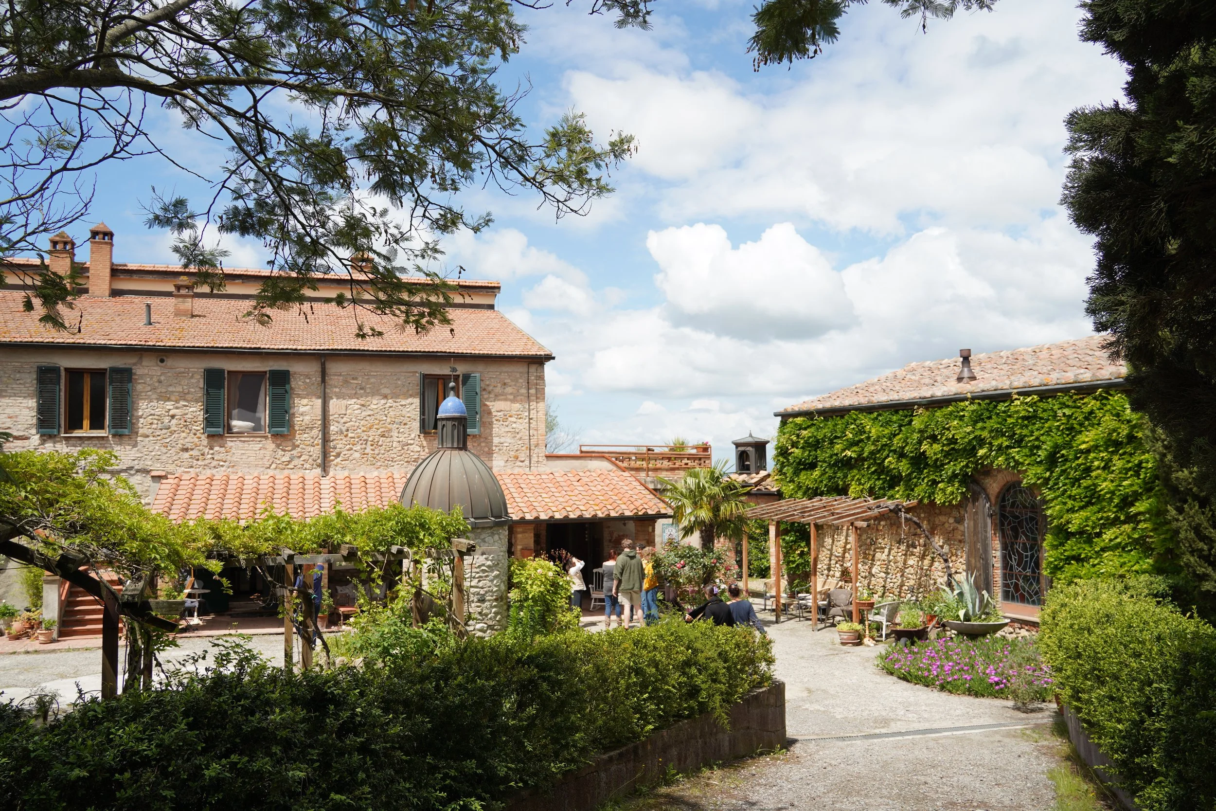 A charming courtyard with stone and brick buildings, lush greenery, flowering plants, and a group of people gathered near an entrance on a partly cloudy day.