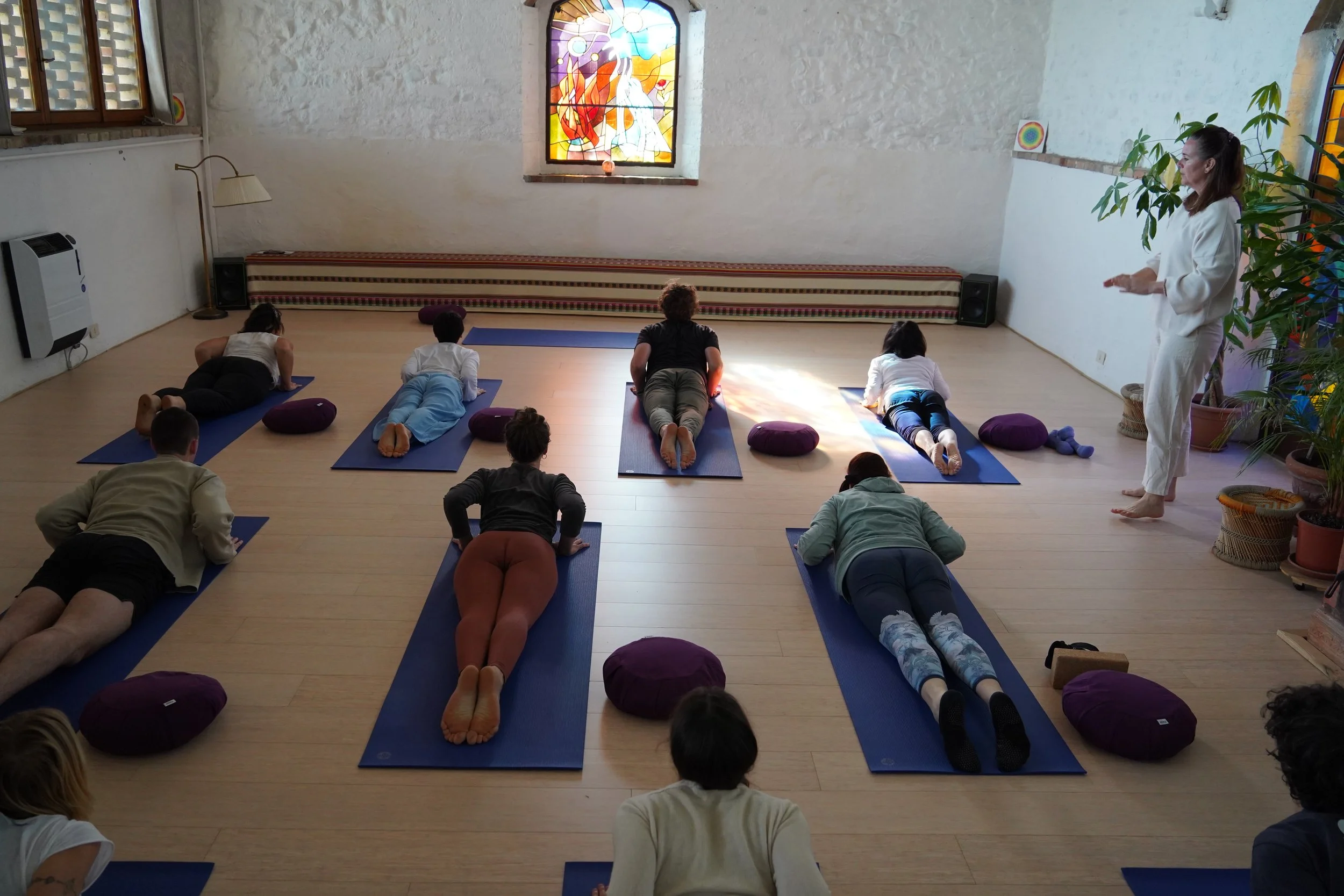 A group of people practicing yoga or meditation in a brightly lit room with wooden floors, cushions, and a stained glass window.