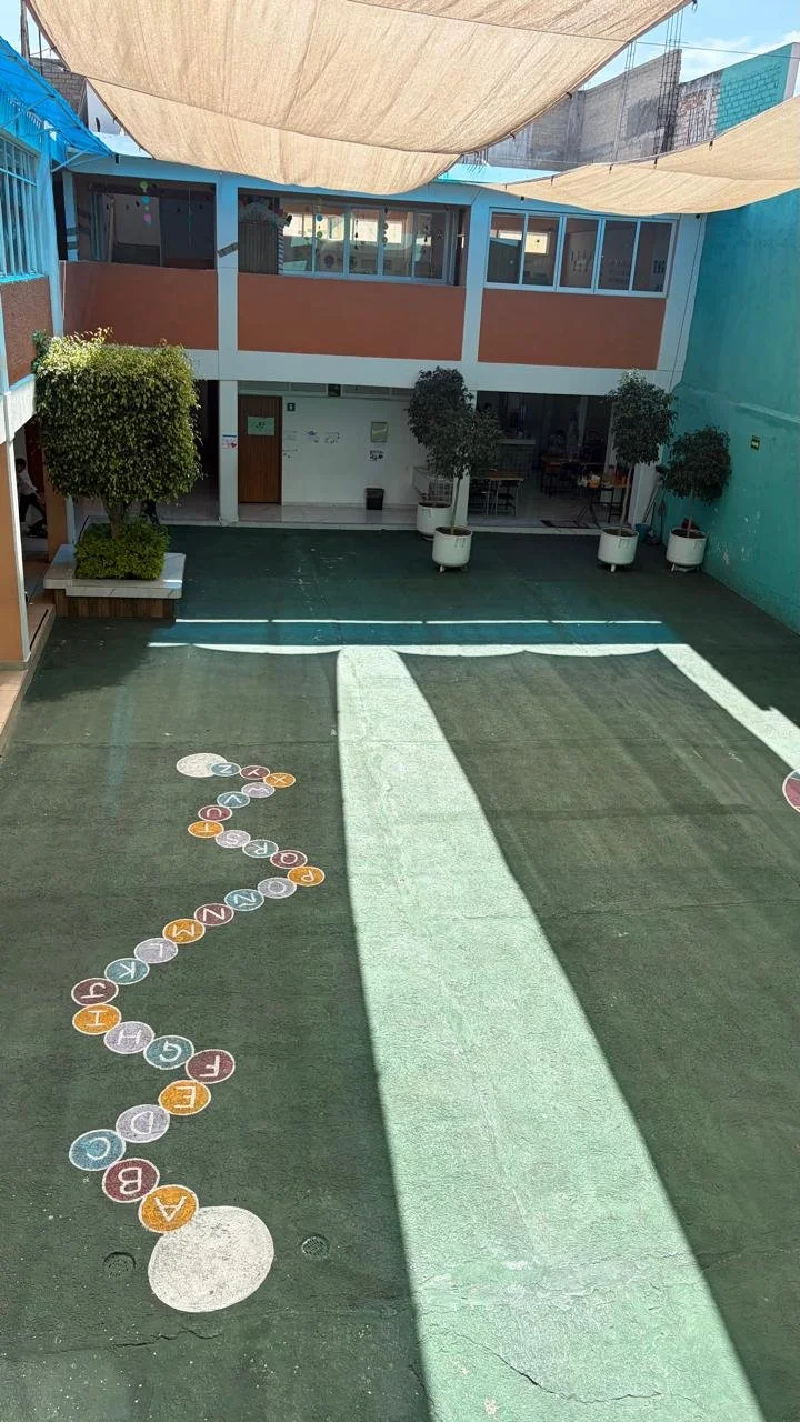 Patio al aire libre con juegos de letras en el suelo formando un camino en zigzag, rodeado de plantas en macetas grandes y una estructura con sombra en la parte superior.