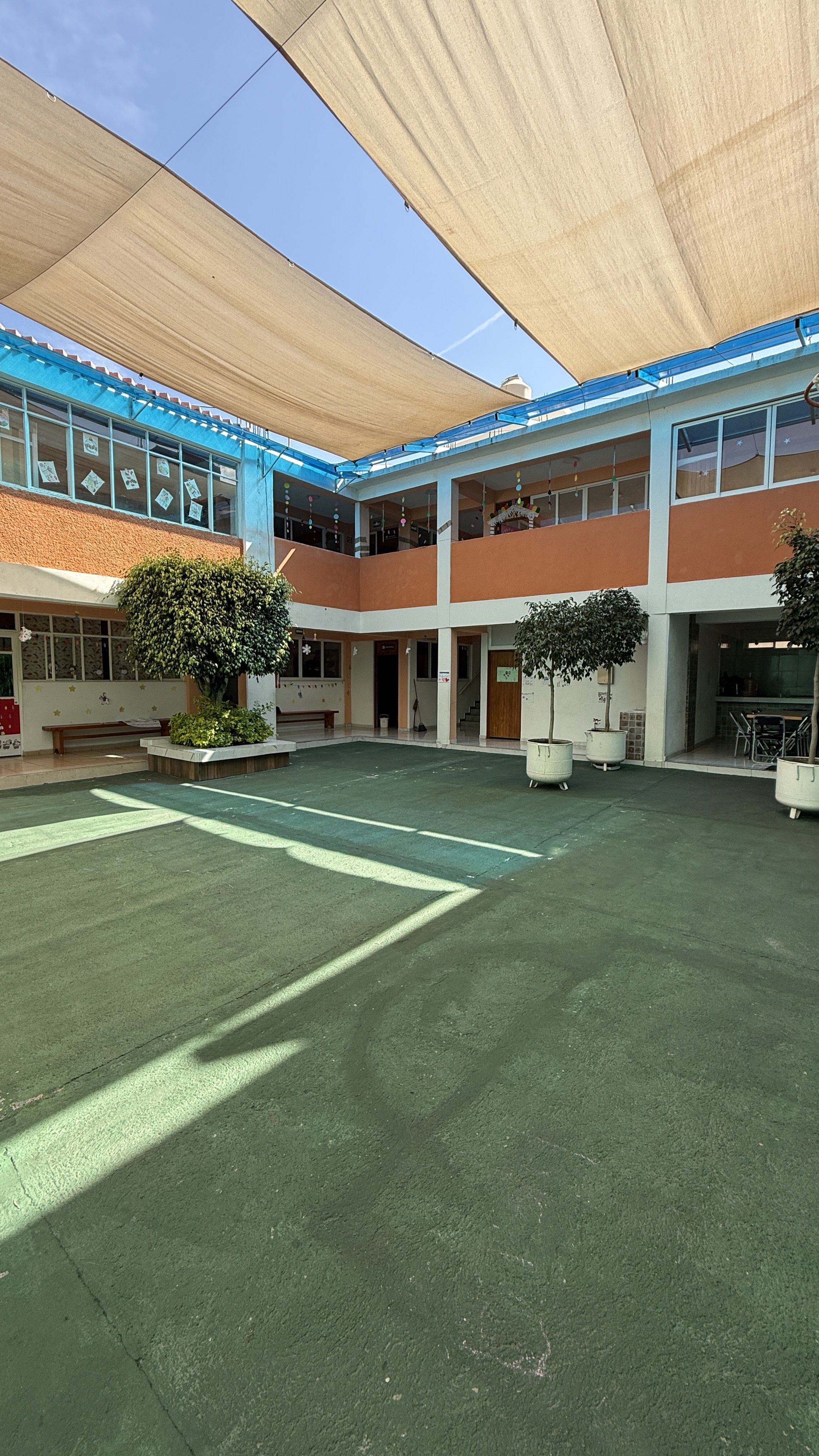 Patio de un colegio con sombra de toldos, paredes color naranja y blancas, plantas en macetas, bancos y ventanas con decoraciones.