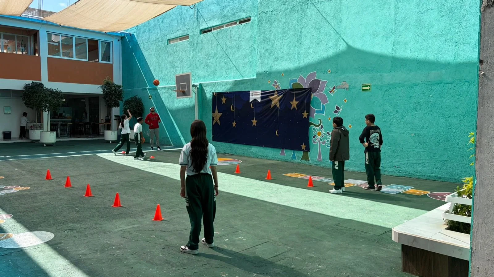 Niños jugando en la cancha de una escuela, algunos lanzando la pelota, otros esperando su turno, en un espacio techado con paredes de color aqua con mural y una lona decorada con estrellas y lunas.