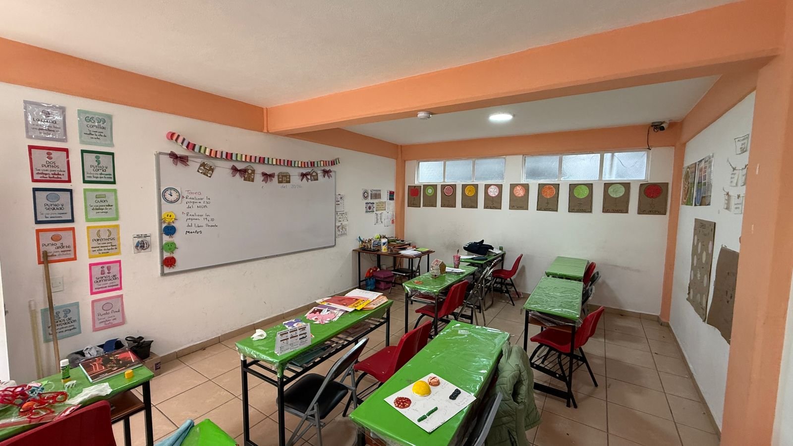 Aula de clases con mesas, sillas rojas, paredes decoradas con carteles y materiales de estudio, y ventanas en la parte superior.