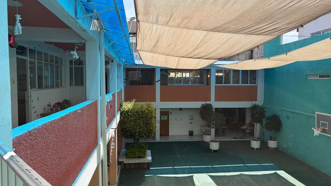 Patio de un colegio con sombra de toldos, paredes de colores vivos, plantas en macetas grandes, y una cancha de básquetbol con aros en los extremos.