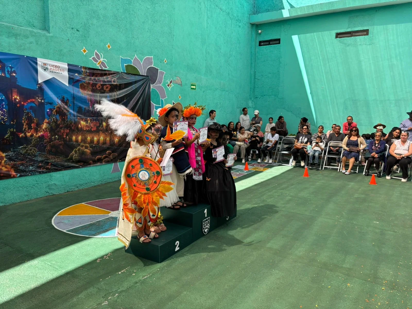 Ceremonia de premiación en un evento cultural con personas vestidas con trajes tradicionales indígenas, en un espacio cerrado decorado con un mural floral y un banner que dice "Liceo Madero Primaria". Hay un público sentado observando el evento.